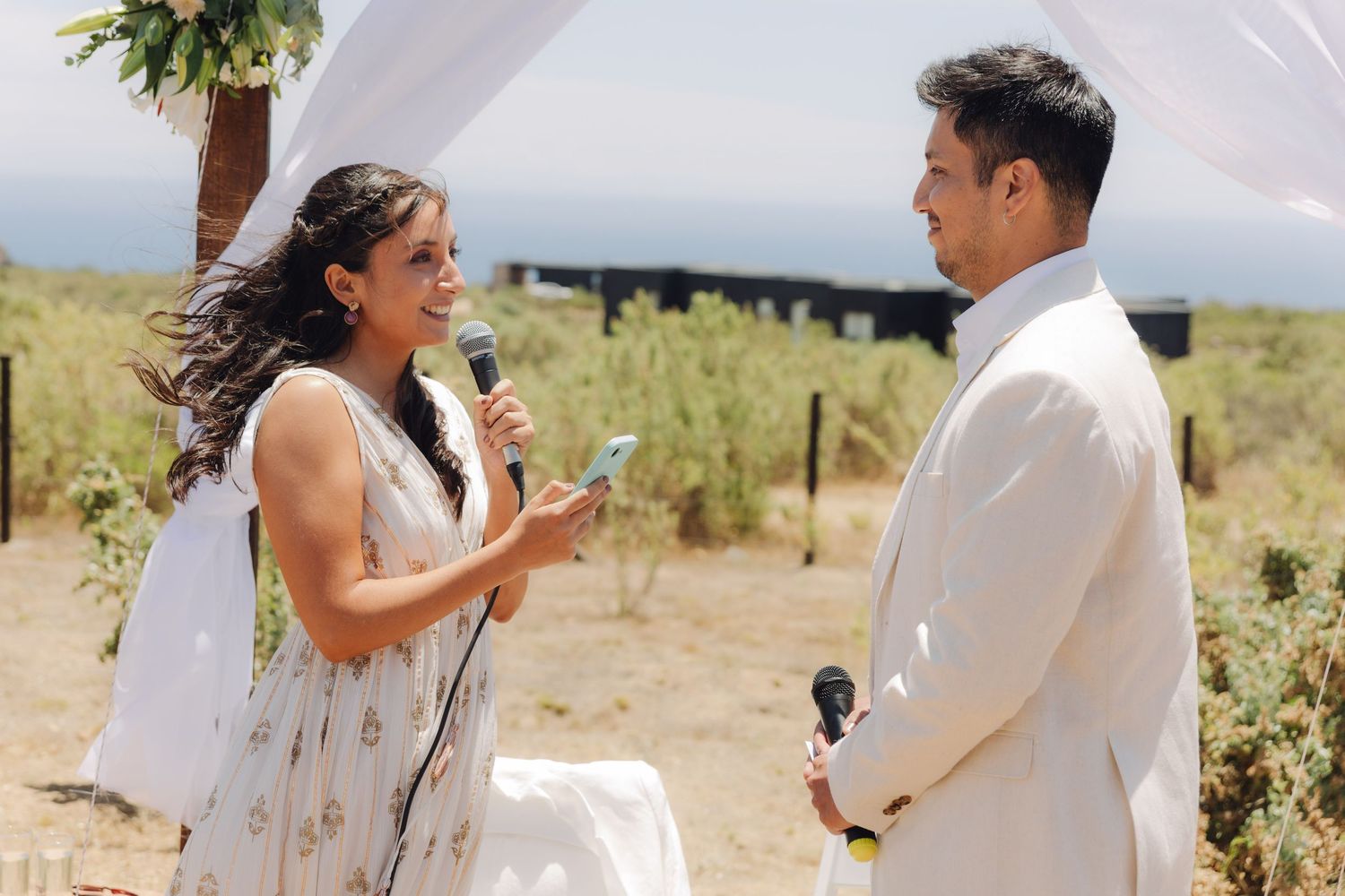 Smiling bride reads vows from phone while groom listens at sunny outdoor wedding ceremony.