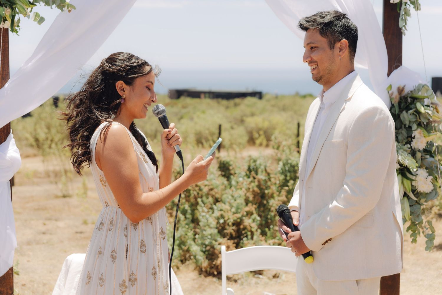 Bride speaks into microphone while exchanging vows with groom under white draped wedding arch outdoors.