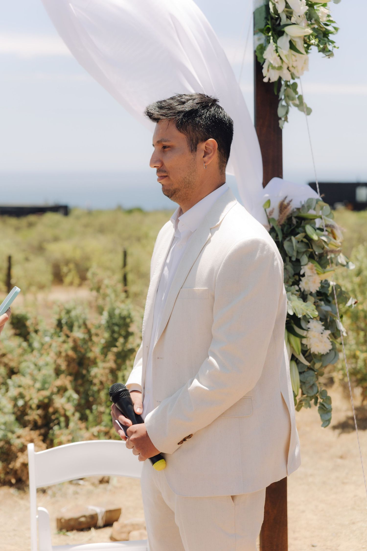 Groom in beige suit holds microphone while standing at outdoor wedding ceremony altar with floral decor.