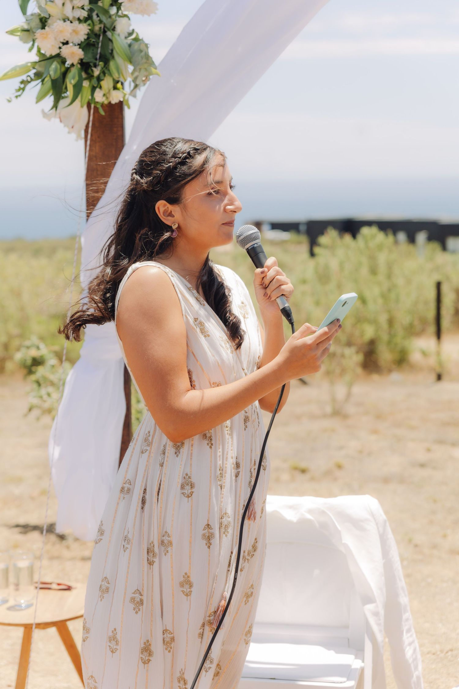 Bride in white floral dress reads vows into microphone at outdoor wedding ceremony with rustic arch.