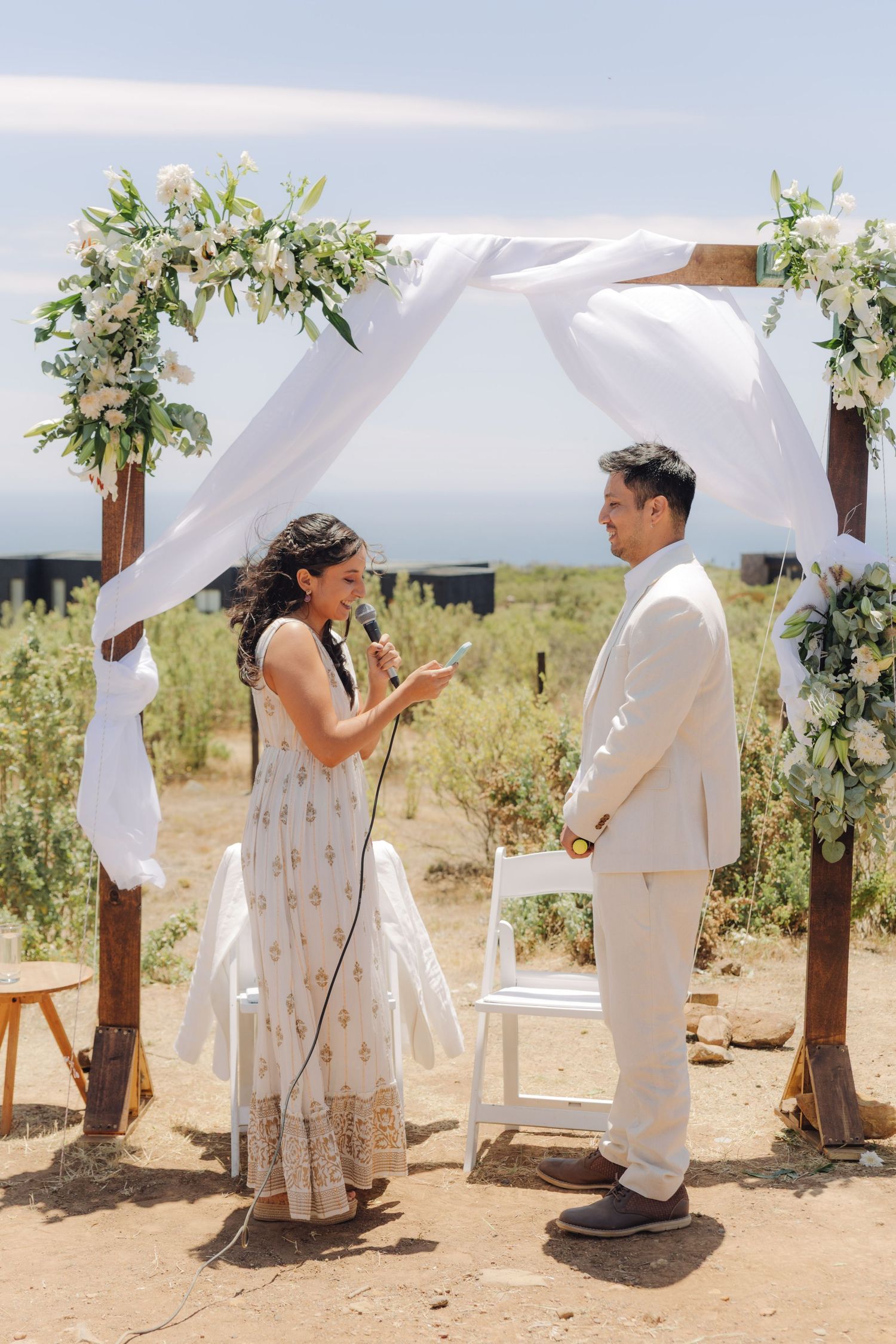 Couple exchanges vows under wooden arch adorned with white draping and white floral arrangements outdoors.