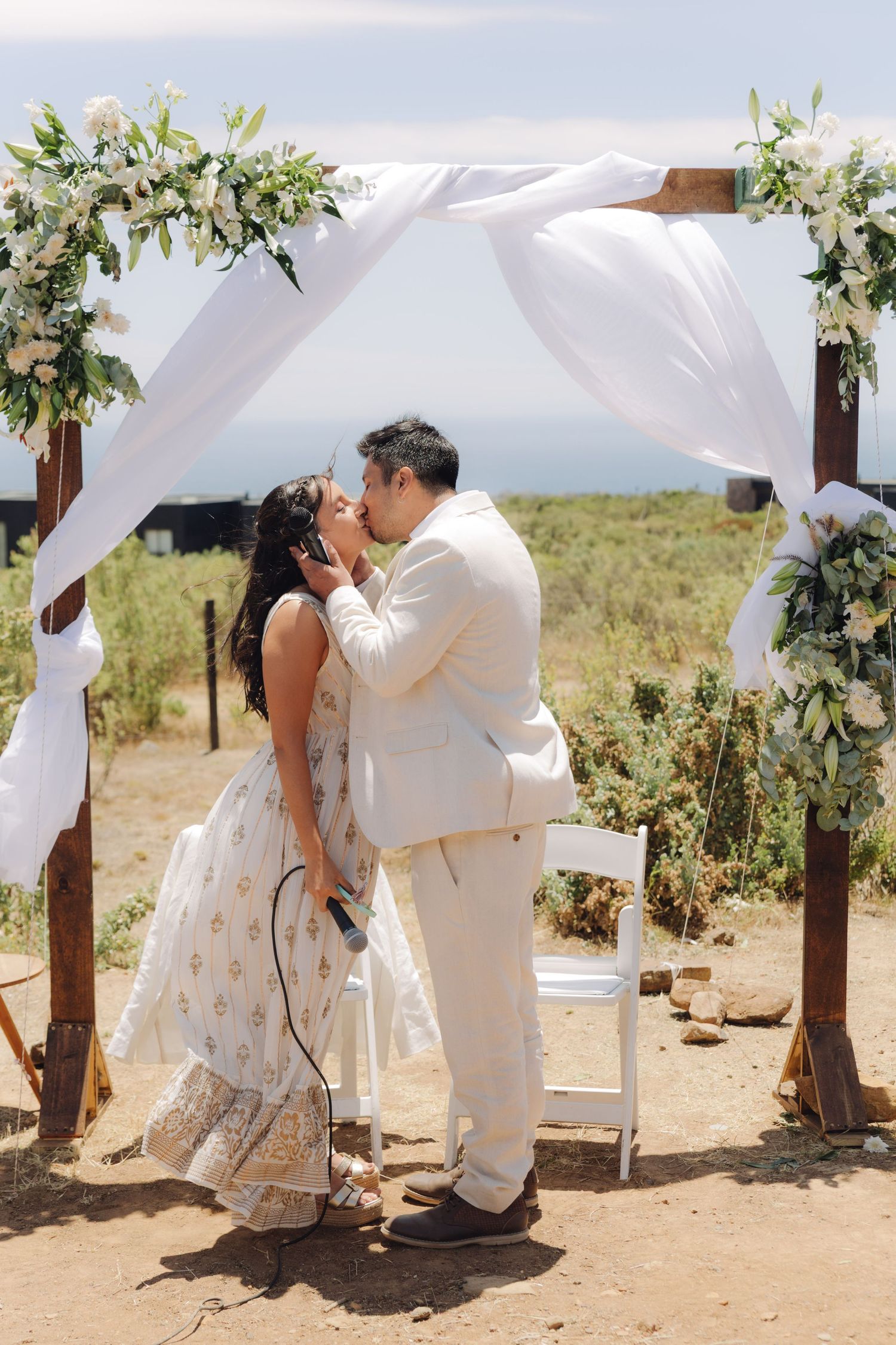 Newlyweds share first kiss under rustic wooden arch with white draping at outdoor wedding ceremony.