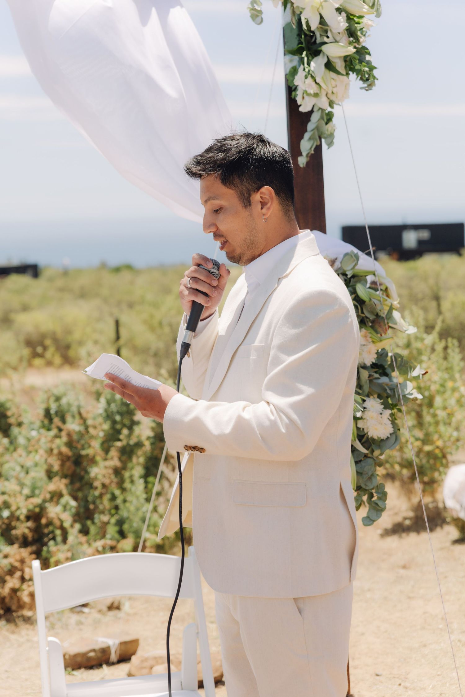 Groom in cream suit reading vows at outdoor wedding ceremony with floral arch and white draping.