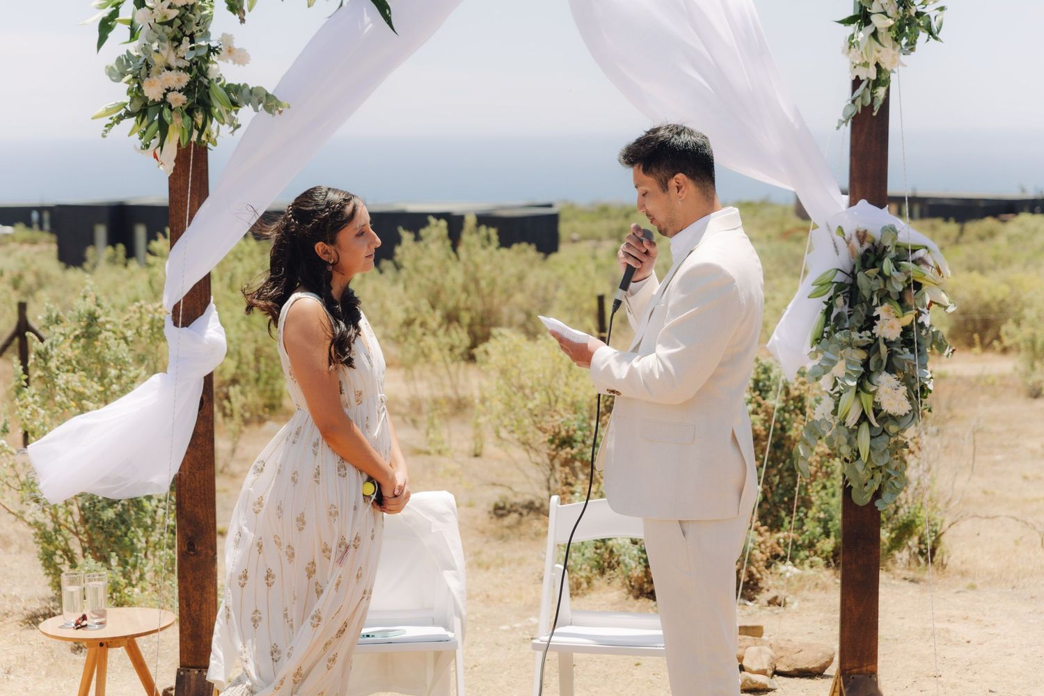Couple exchanging vows under a wooden arch with white draping and greenery at outdoor wedding.