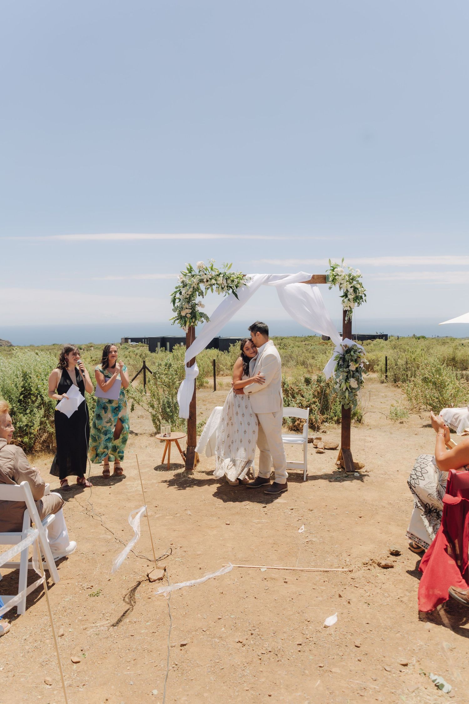 Bride and groom share first kiss under floral arch at sunny outdoor wedding ceremony with guests.