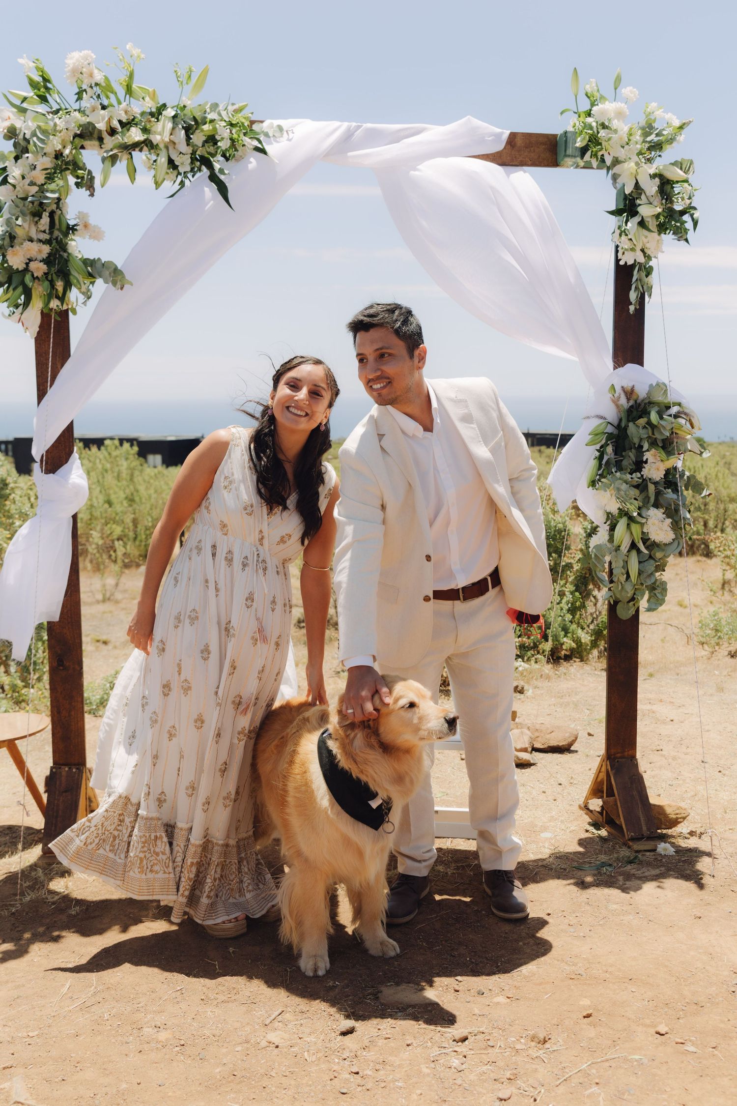 Newlyweds pose with golden retriever dog under decorated wedding arch at outdoor ceremony.