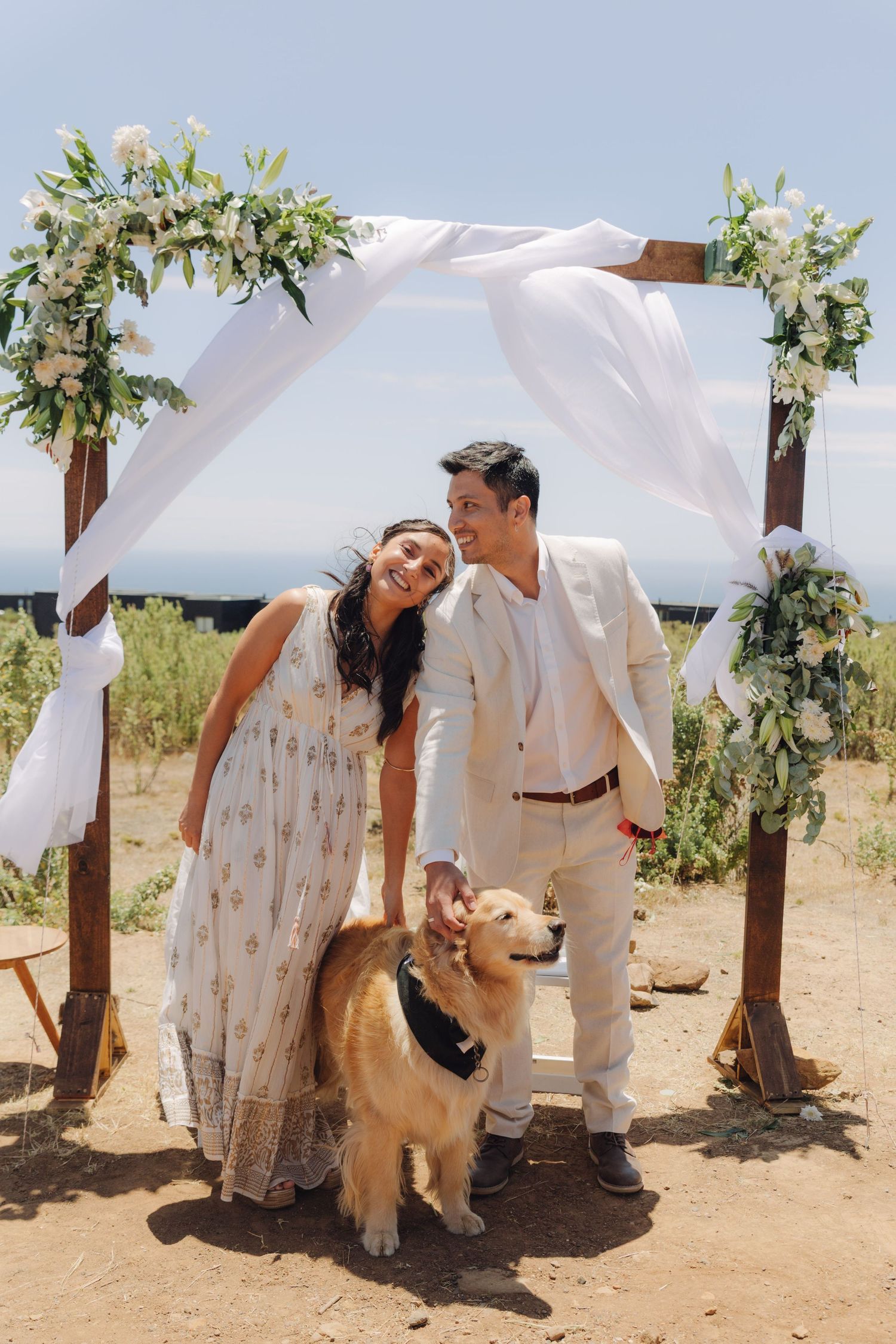 Smiling bride and groom stand with their golden retriever under a floral wedding arch outdoors.
