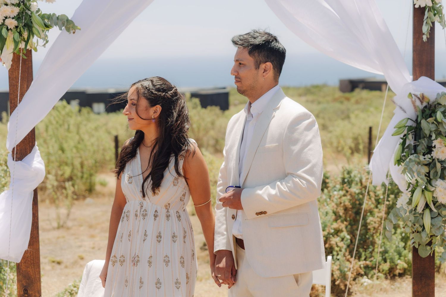 Bride and groom hold hands under white draped wooden arch at scenic outdoor coastal wedding.