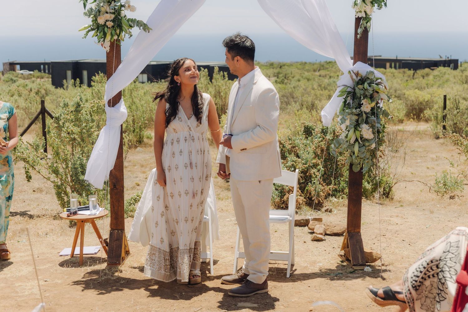 Bride and groom smile at each other under a flower-adorned arch at an outdoor wedding ceremony.
