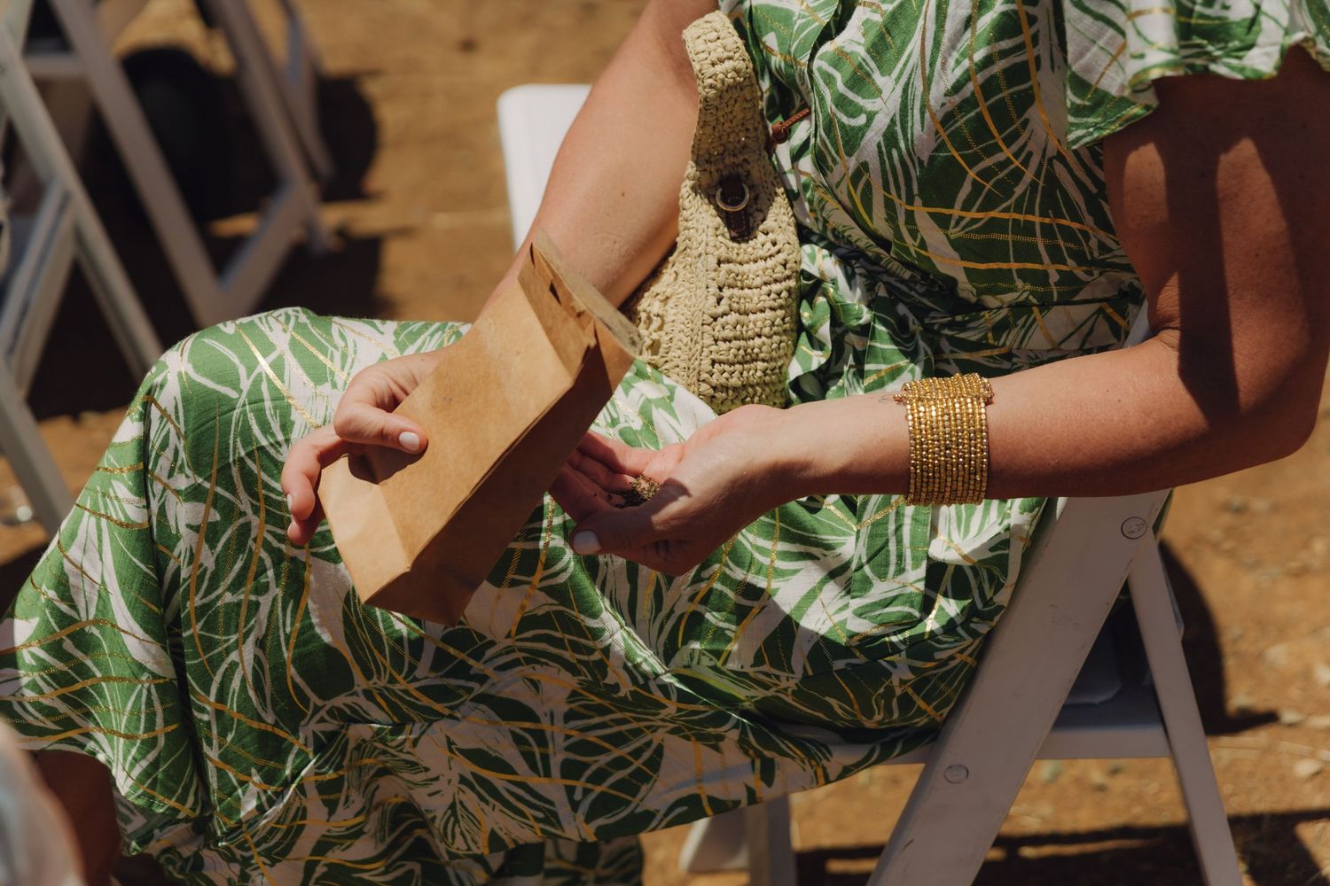 Wedding guest in green floral dress holds brown paper bag while seated at outdoor ceremony.