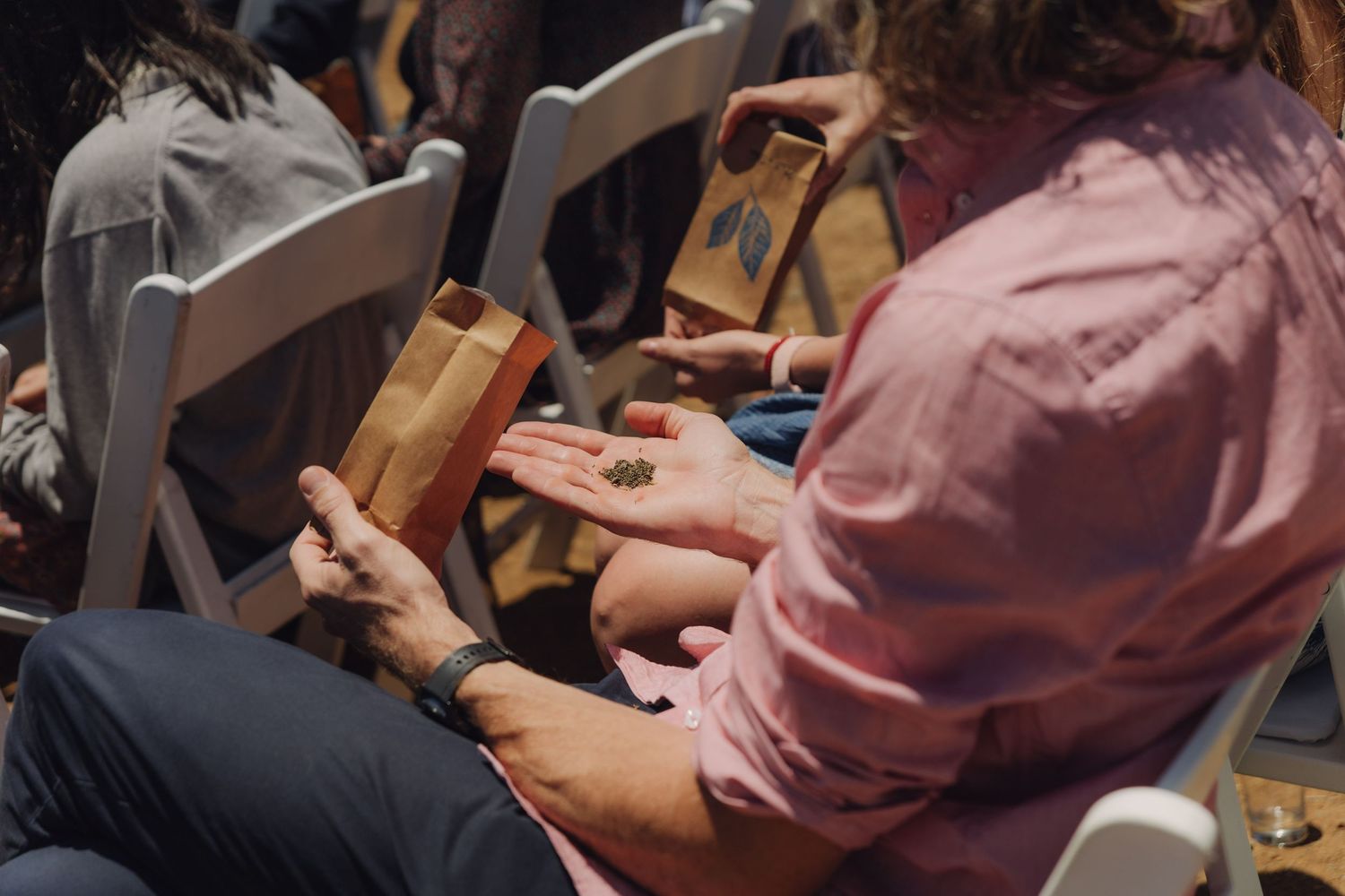 Wedding guests hold small brown paper bags with confetti at an outdoor ceremony celebration.