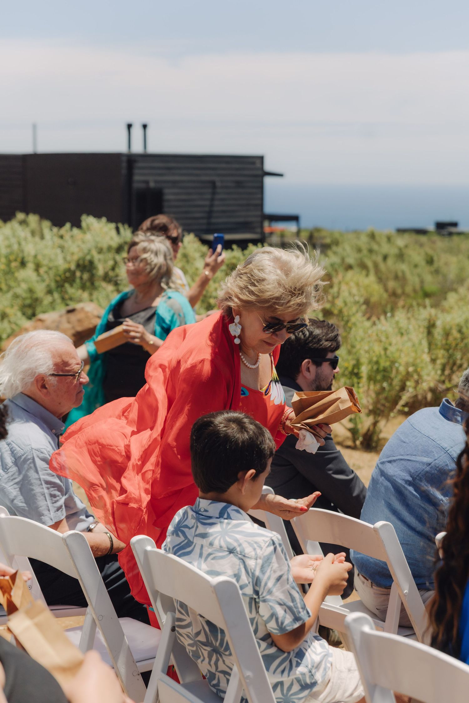 Wedding guests seated outdoors, woman in red outfit interacts with child at sunny ceremony.