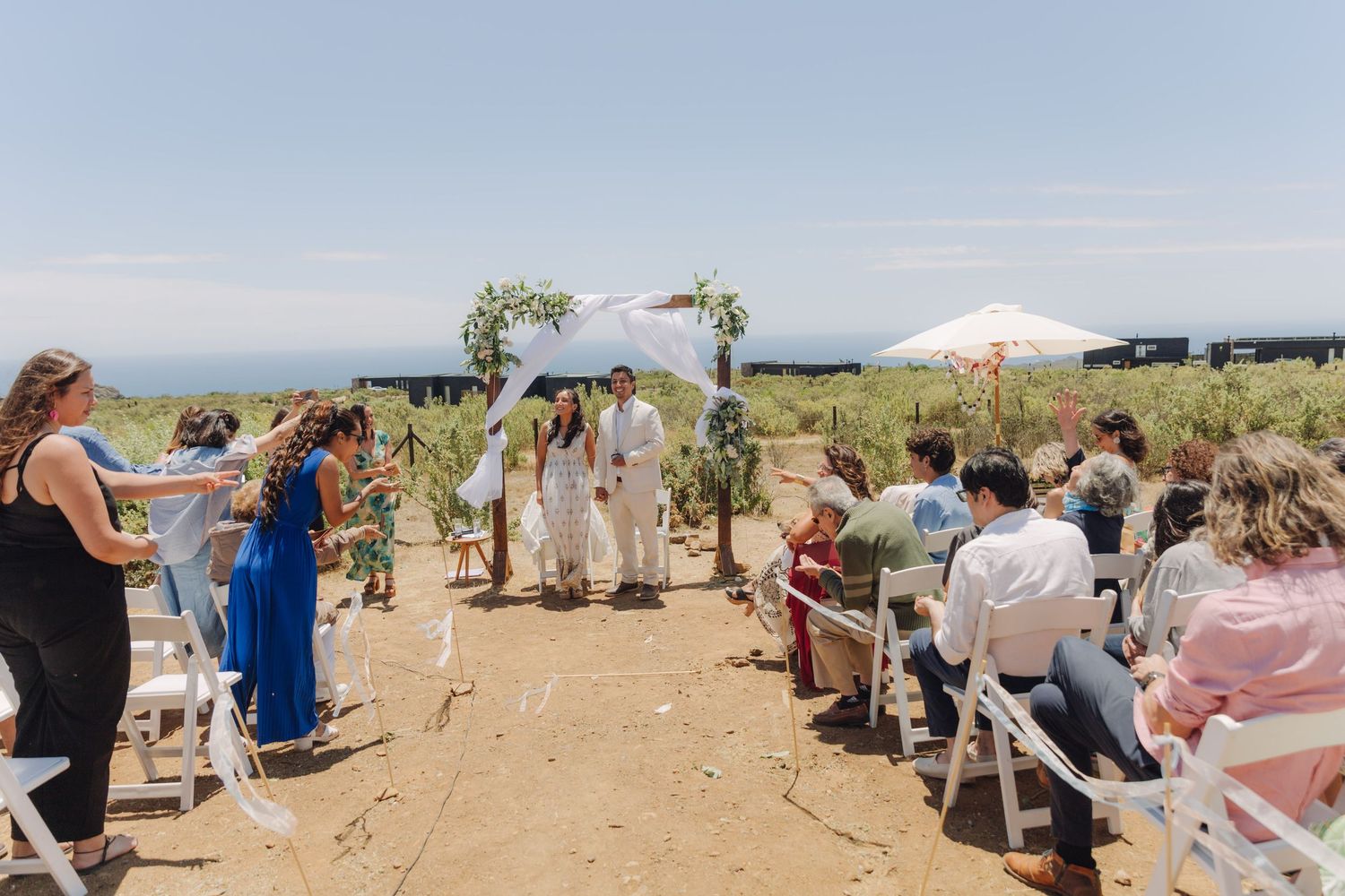 Outdoor wedding ceremony with guests seated on white chairs as couple exchanges vows under a floral arch.