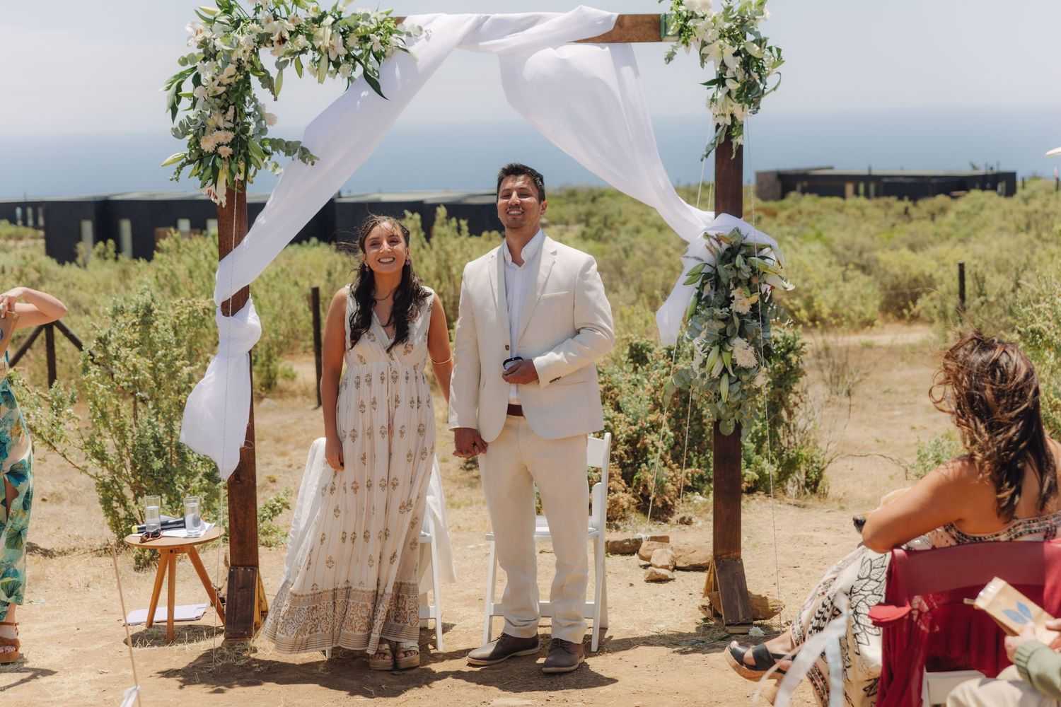 Bride and groom stand together smiling under a white draped wooden arch decorated with greenery outdoors.