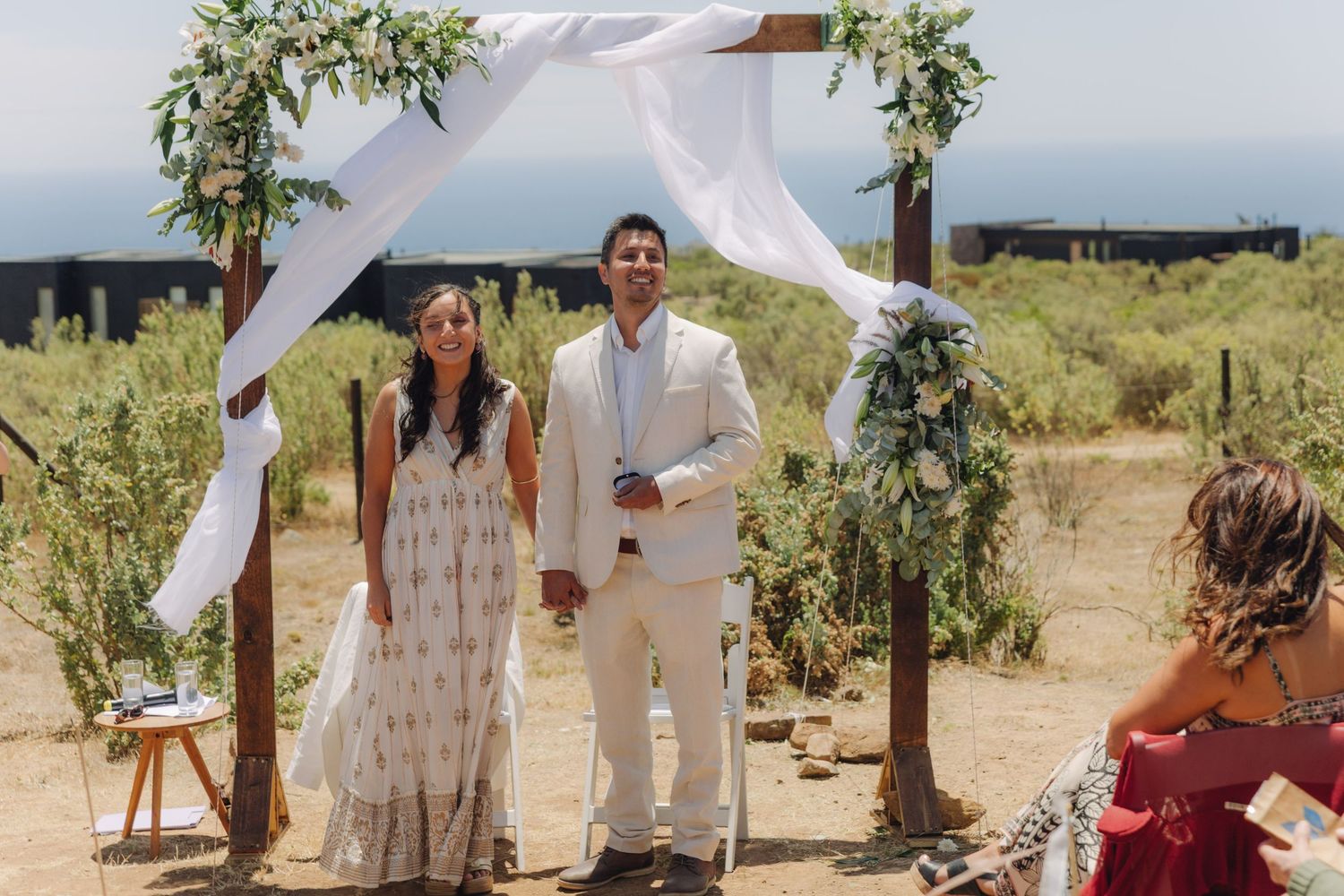 Newlyweds pose under a rustic wooden wedding arch adorned with white fabric and lush greenery in a sunny outdoor setting.