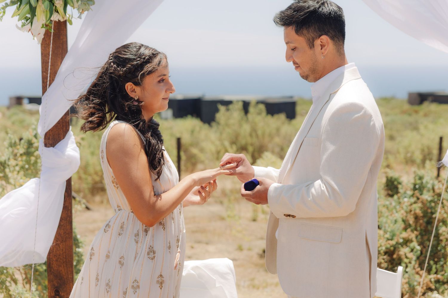 Groom places wedding ring on brides finger during outdoor ceremony beneath a floral decorated wooden arch.