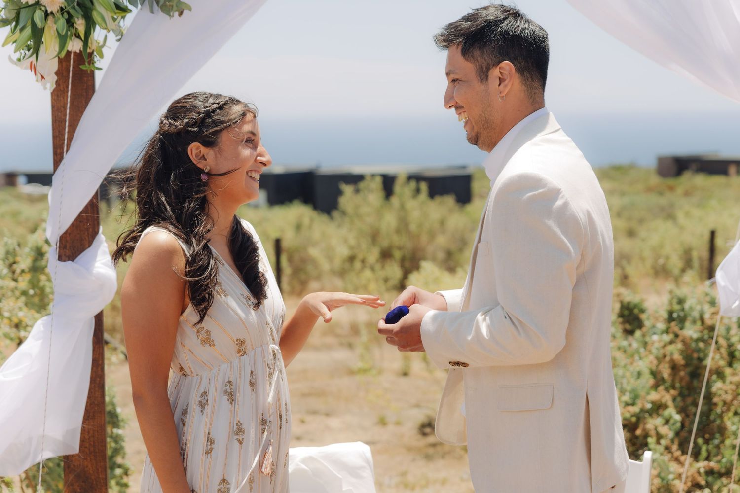 Bride laughs joyfully as groom places wedding ring on her finger during sunny outdoor ceremony.