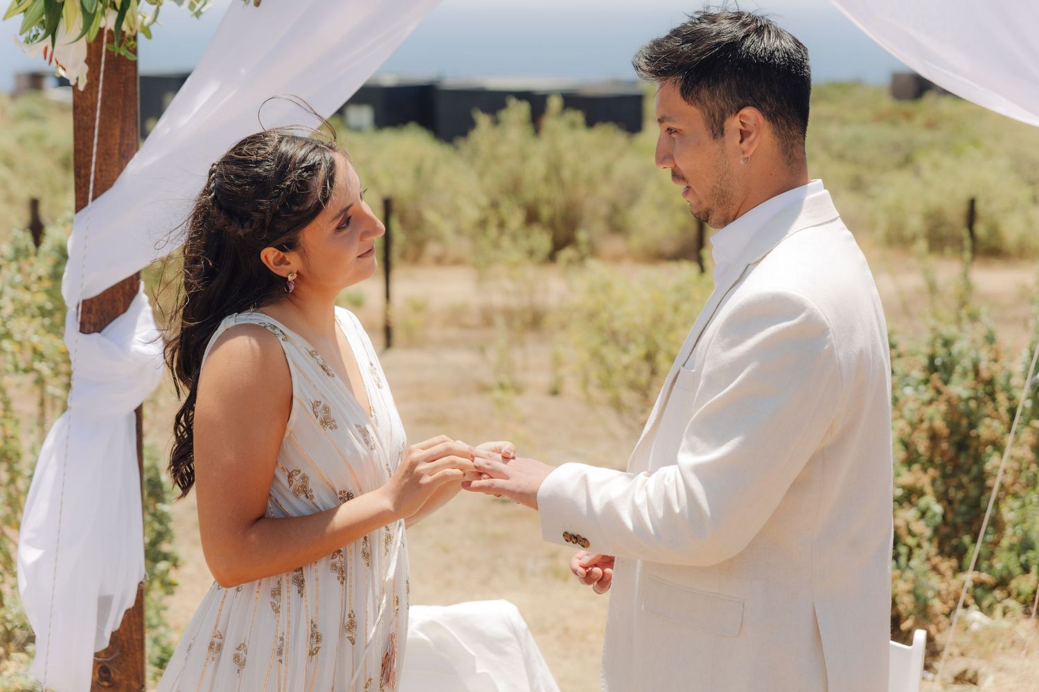 Couple exchanges rings during an intimate outdoor wedding ceremony beneath a white draped wooden arch.