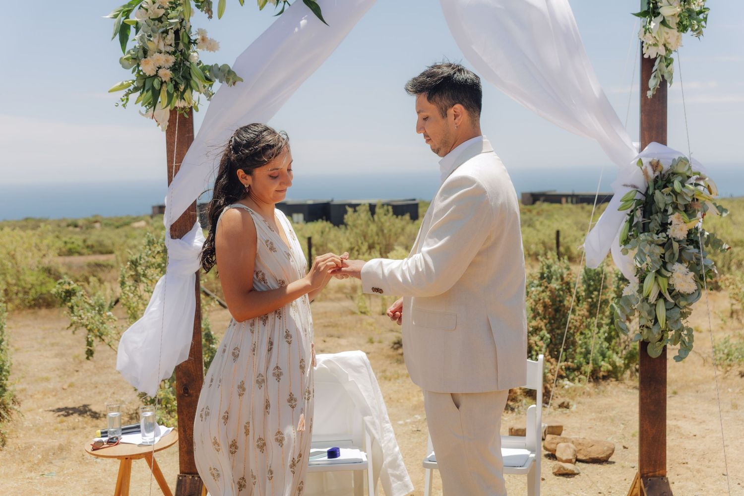 Couple exchanges wedding rings under a wooden arch with white draping and floral greenery in a sunny outdoor setting.