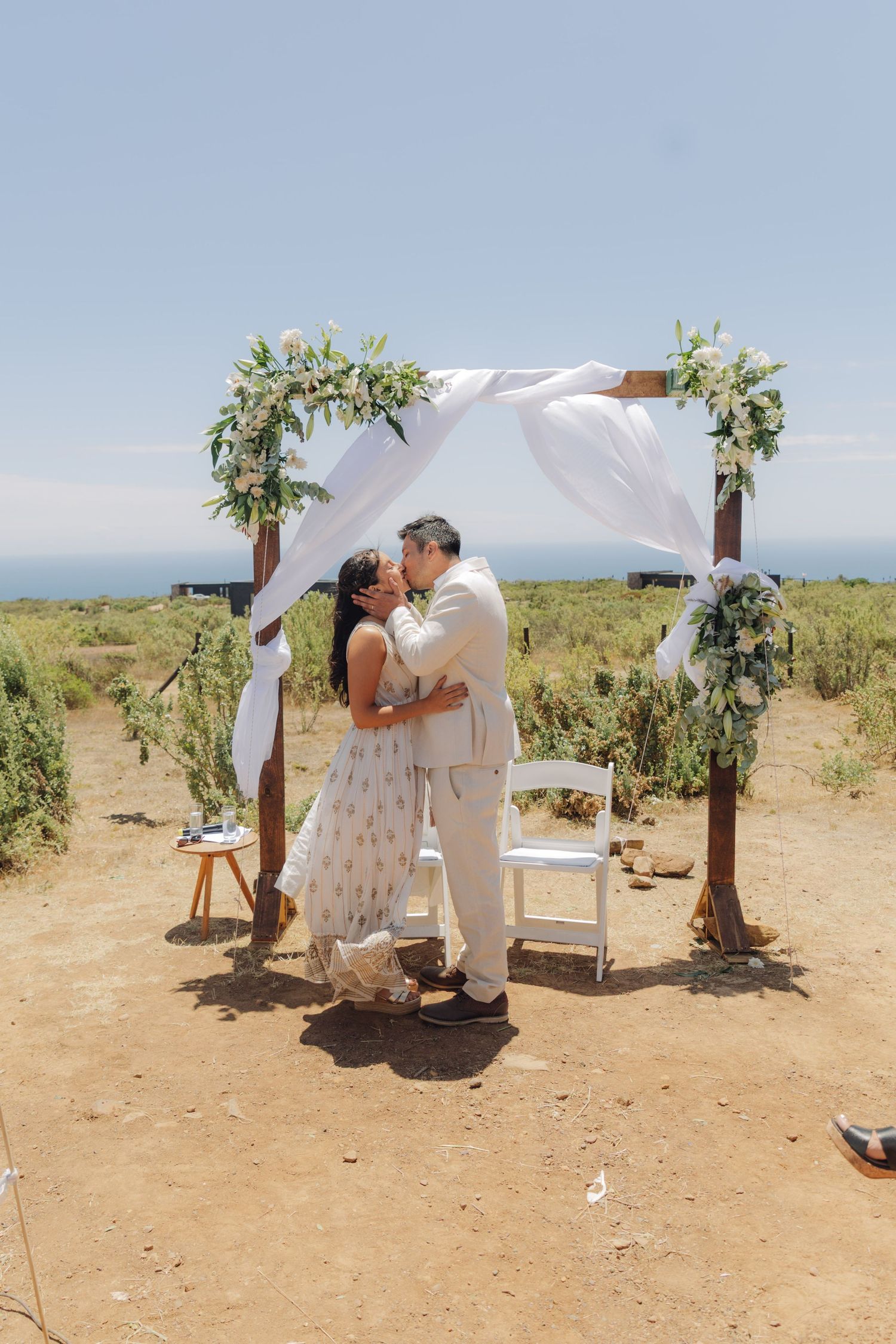 Newlyweds share their first kiss under a white draped wooden wedding arch decorated with lush greenery outdoors.