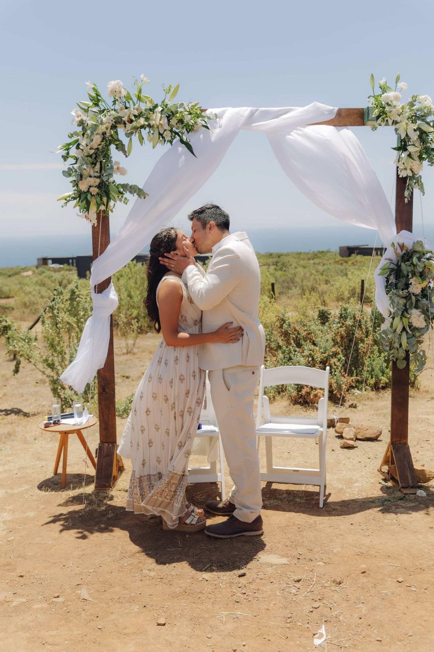 Couple shares first kiss under a floral wooden arch with white draping at an outdoor wedding ceremony.