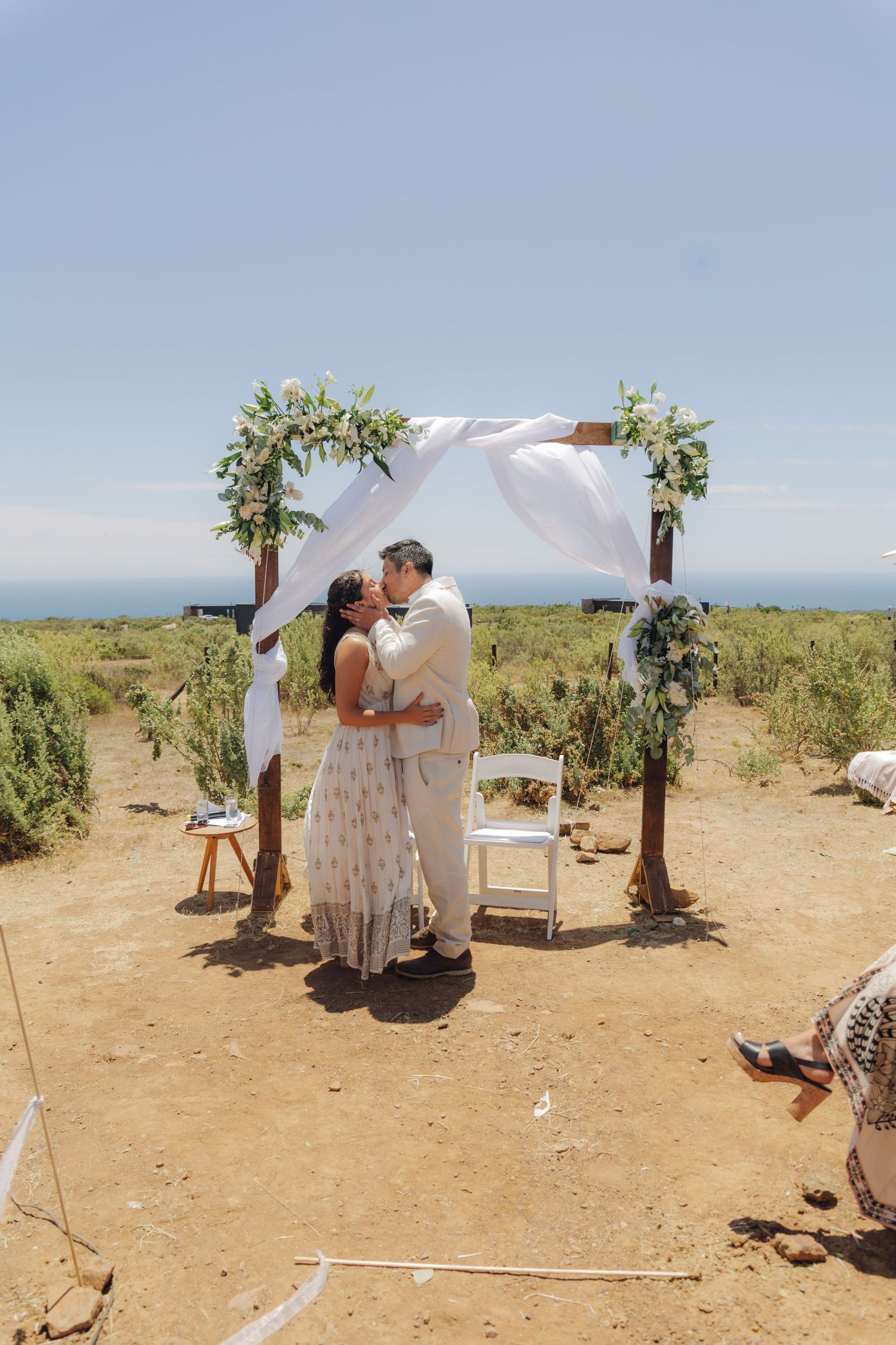 Newlyweds kiss beneath a rustic wooden arch adorned with greenery and white fabric at a coastal wedding.