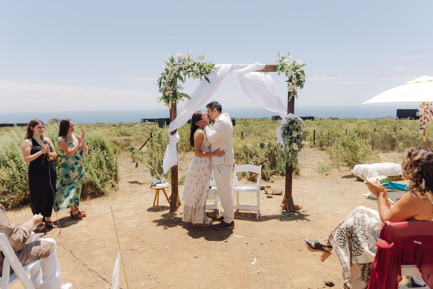 Wedding couple kisses under a decorated arch surrounded by seated guests at an outdoor seaside ceremony.