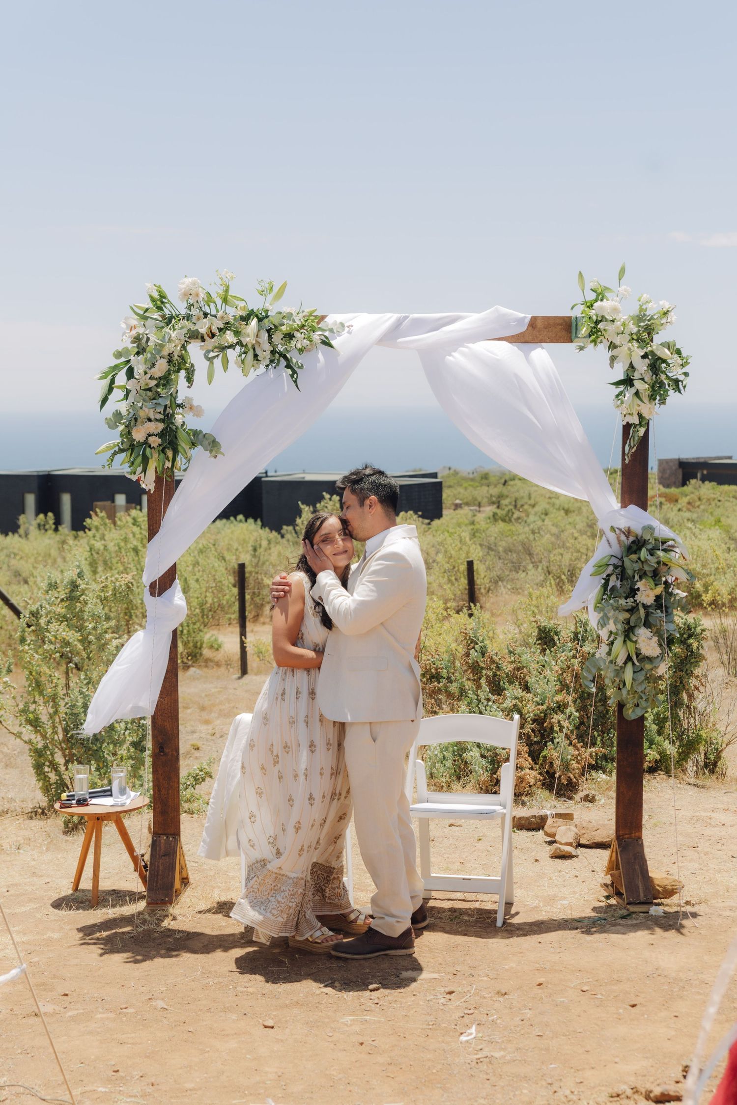Bride and groom embrace tenderly under a white-draped wooden arch with floral arrangements at their wedding.