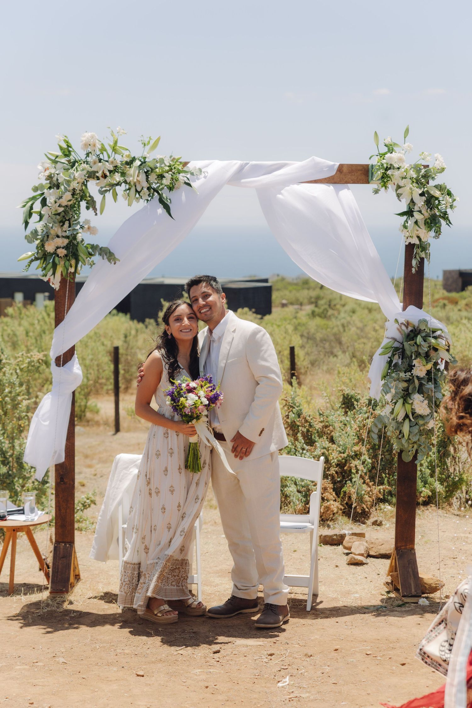 Smiling newlyweds pose under a floral wooden arch, bride holding a colorful bouquet at outdoor wedding.