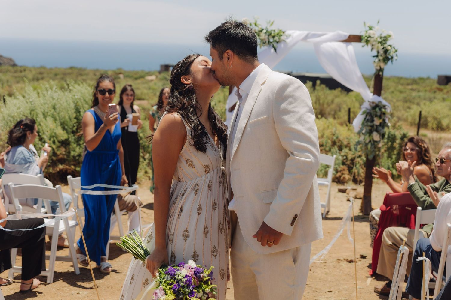 Newlyweds share a passionate kiss while guests celebrate at their outdoor coastal wedding ceremony.