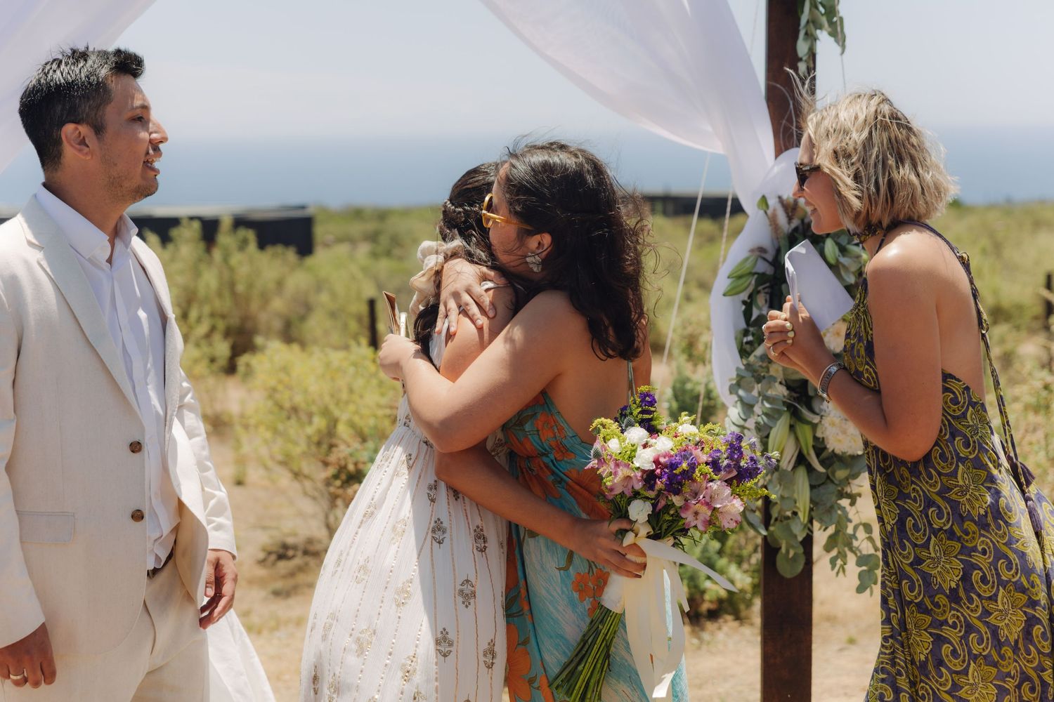 Bride shares a warm hug with a guest in a sari at an outdoor cliffside wedding ceremony.