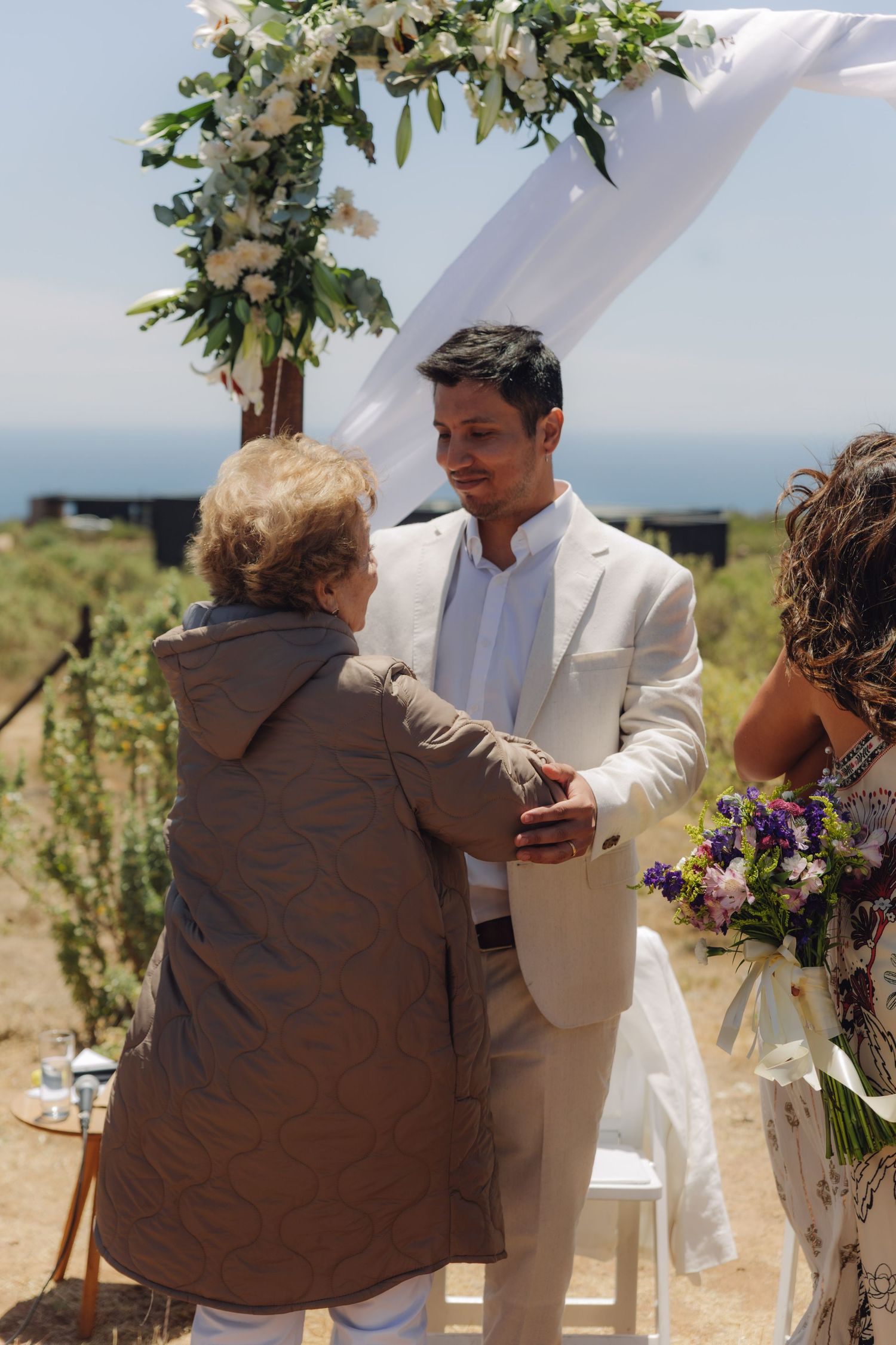 Groom in beige suit warmly greets an elderly guest under a floral arch at an outdoor wedding.