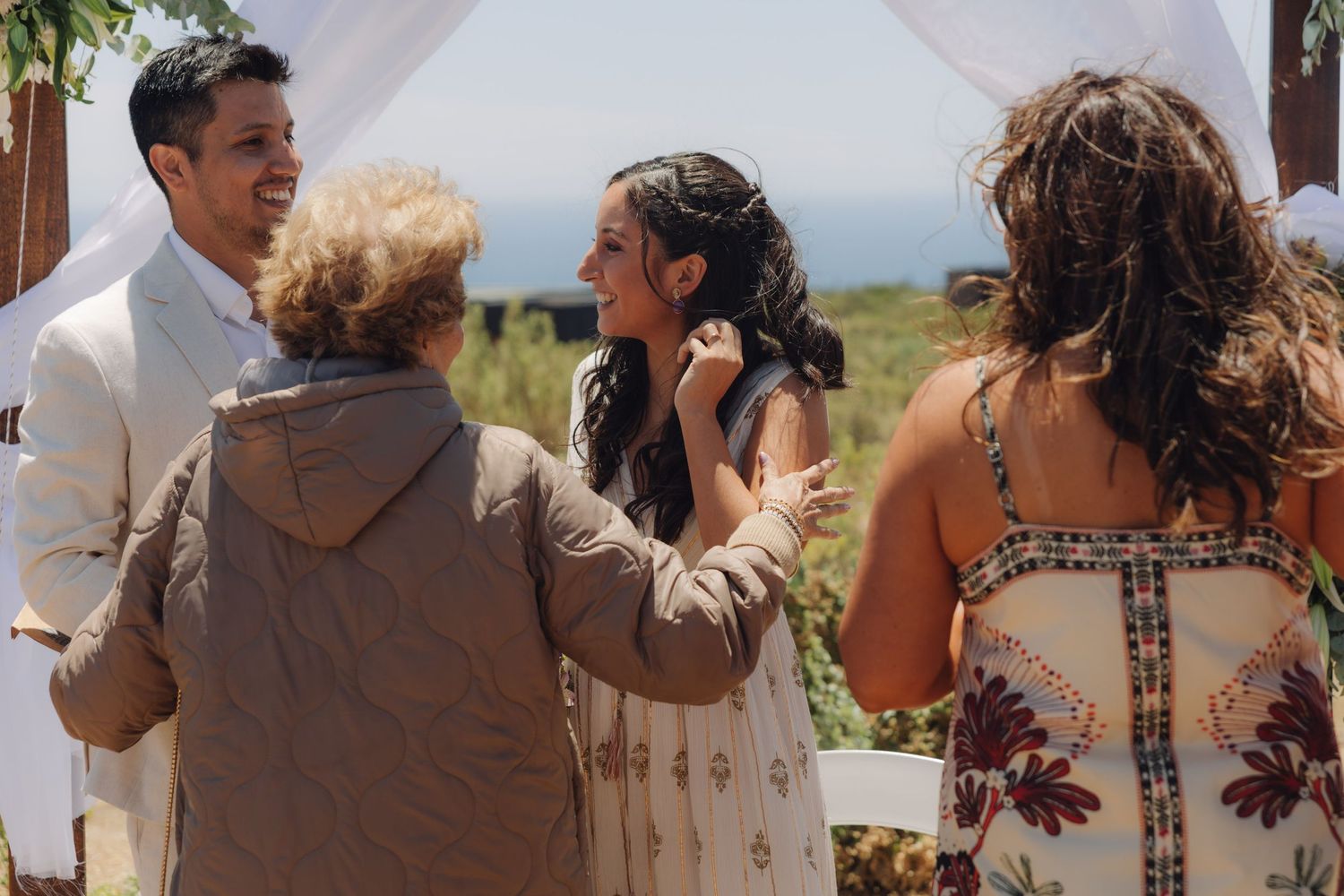Smiling bride receives a tender touch from an elderly guest at a sunny outdoor wedding ceremony.