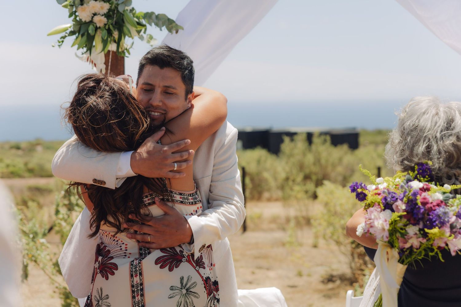 Groom shares a heartfelt embrace with a guest in a floral dress at an outdoor cliffside wedding.