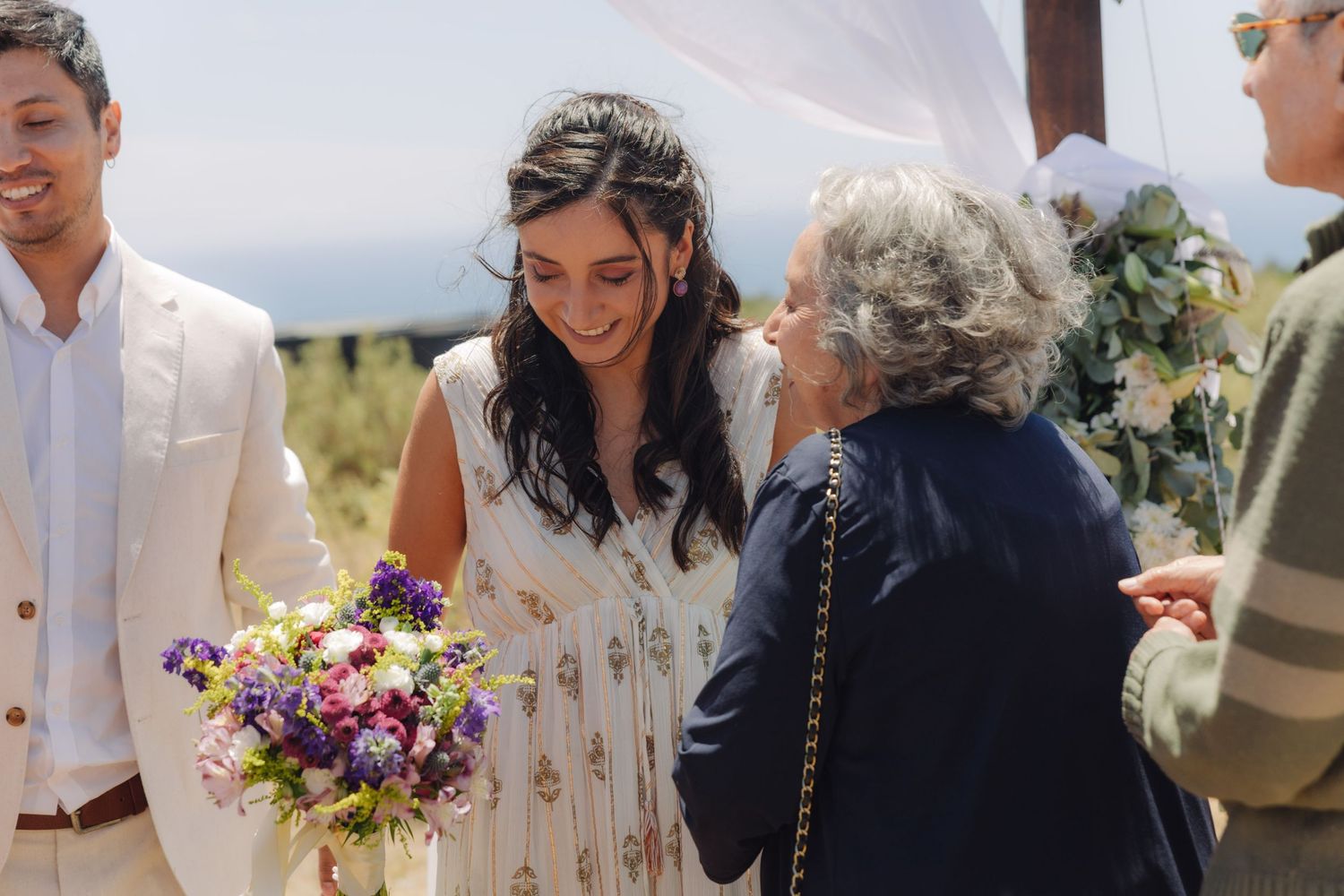 Radiant bride holding purple bouquet laughs with an elderly guest at a sunny outdoor wedding.