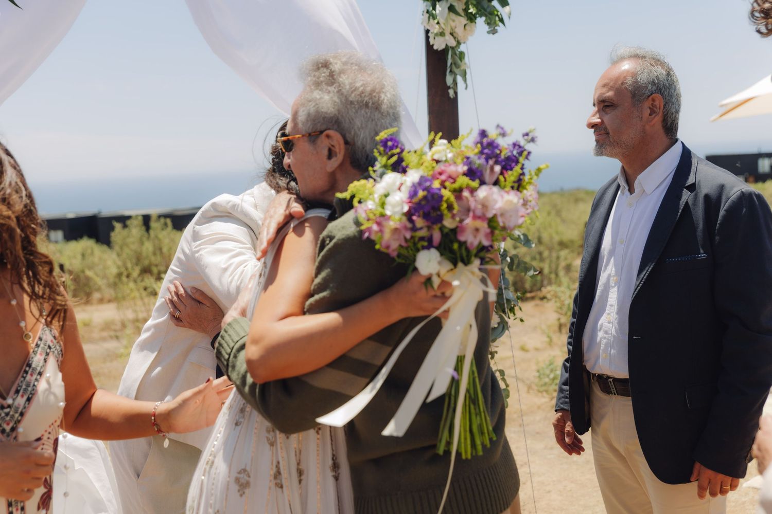 Bride hugs an elderly guest warmly while holding a colorful bouquet at a sunny outdoor wedding.