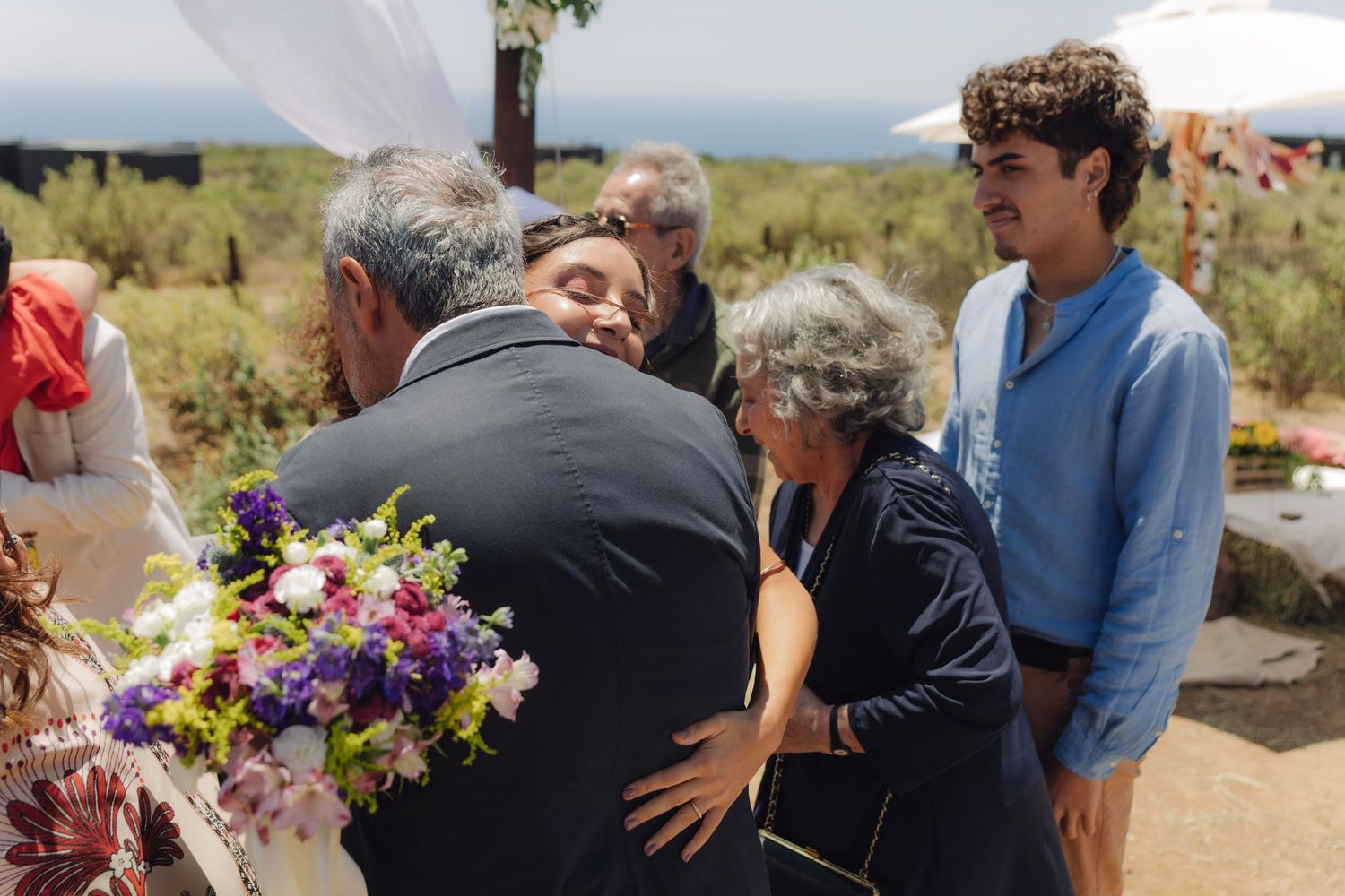 Guests embracing at an outdoor wedding ceremony with colorful bouquet and ocean view.