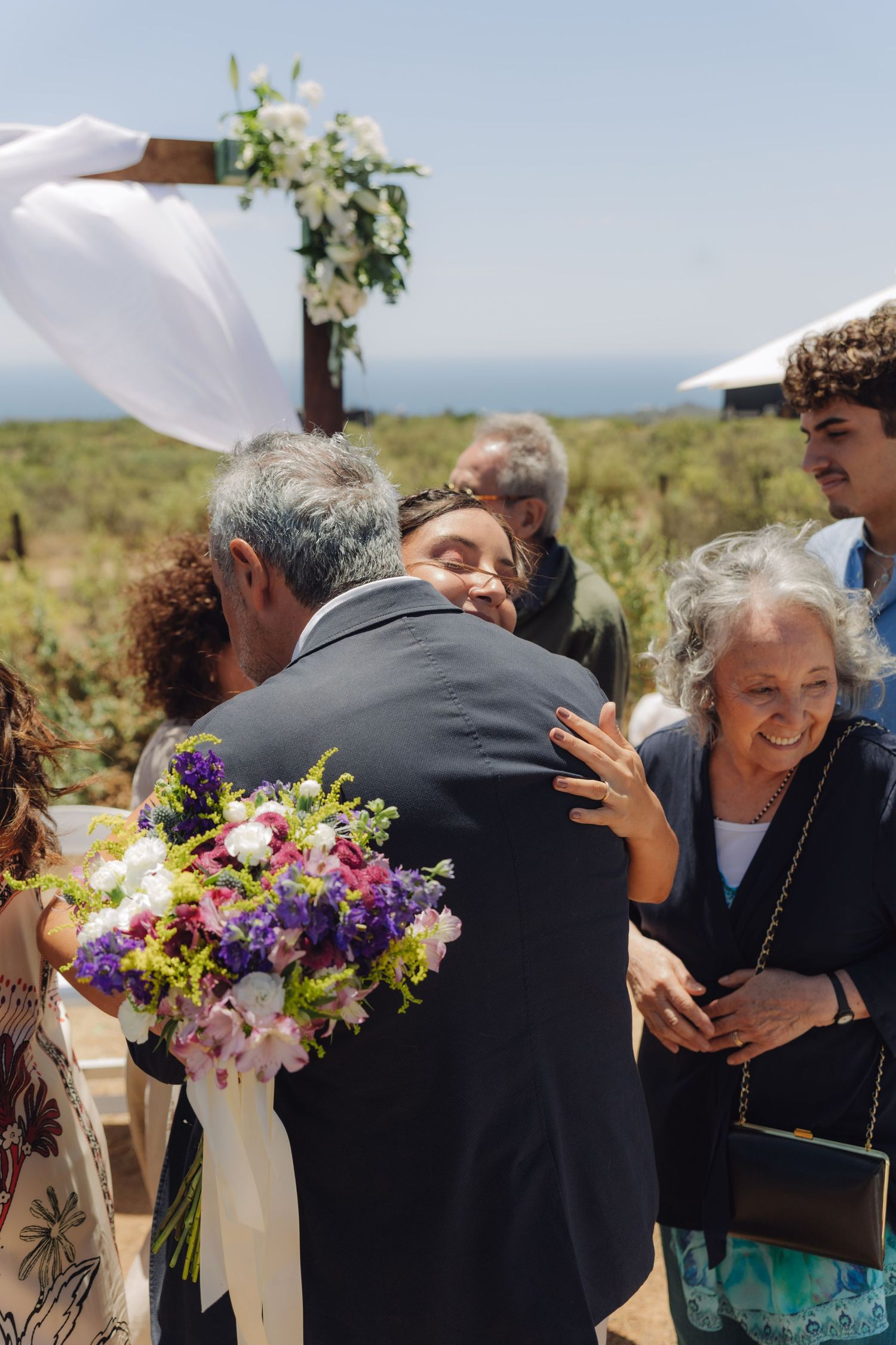 Bride hugging guest at sunny outdoor wedding ceremony with floral arch and bouquet.