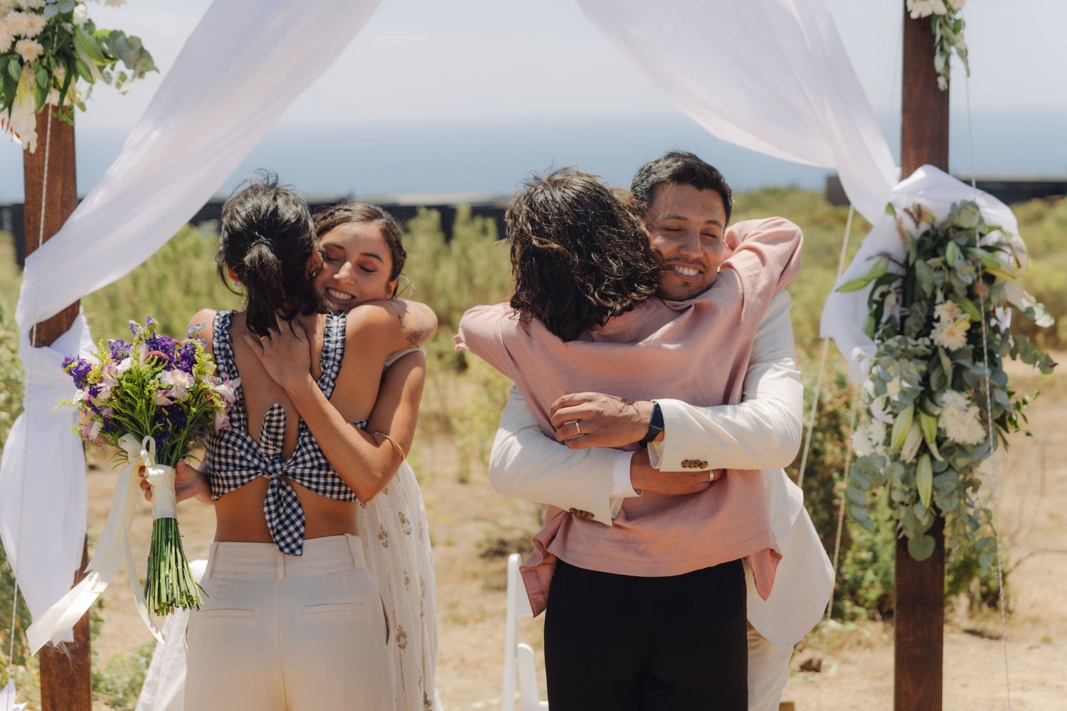 Two couples sharing warm embraces at an outdoor wedding ceremony under a wooden arch.