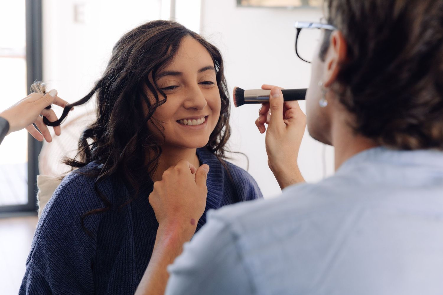 Woman smiling during bridal makeup session with stylist applying cosmetics indoors.