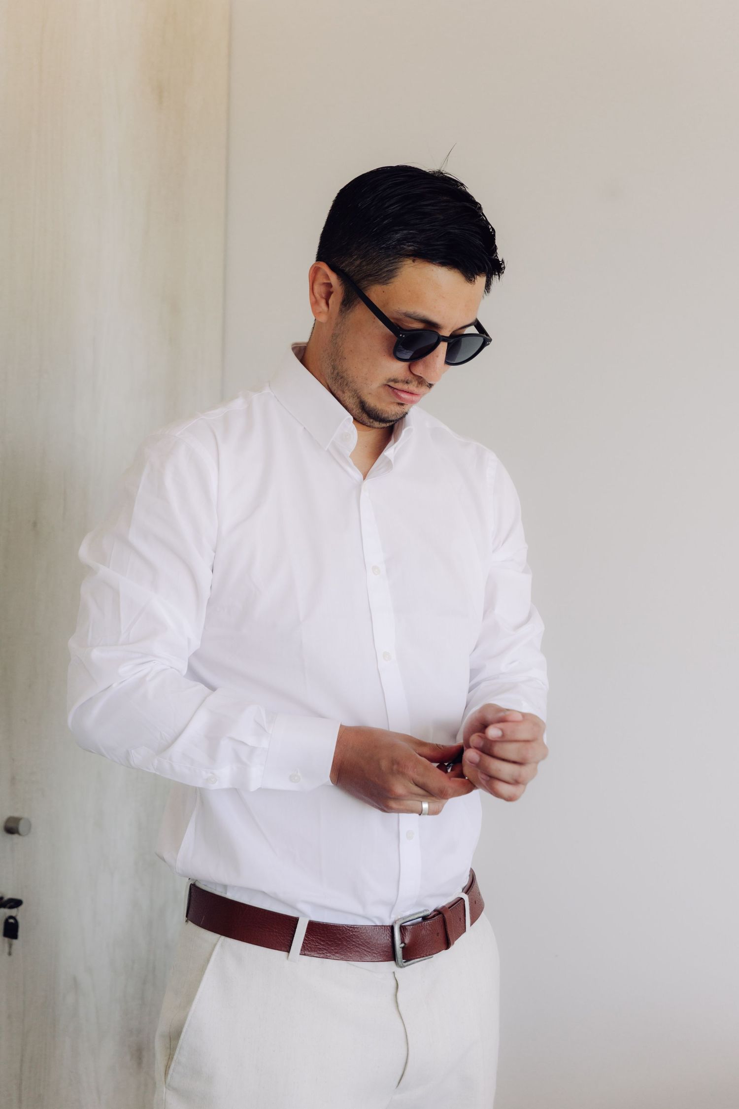 Stylish groom in white shirt and sunglasses adjusting cufflinks before wedding ceremony.