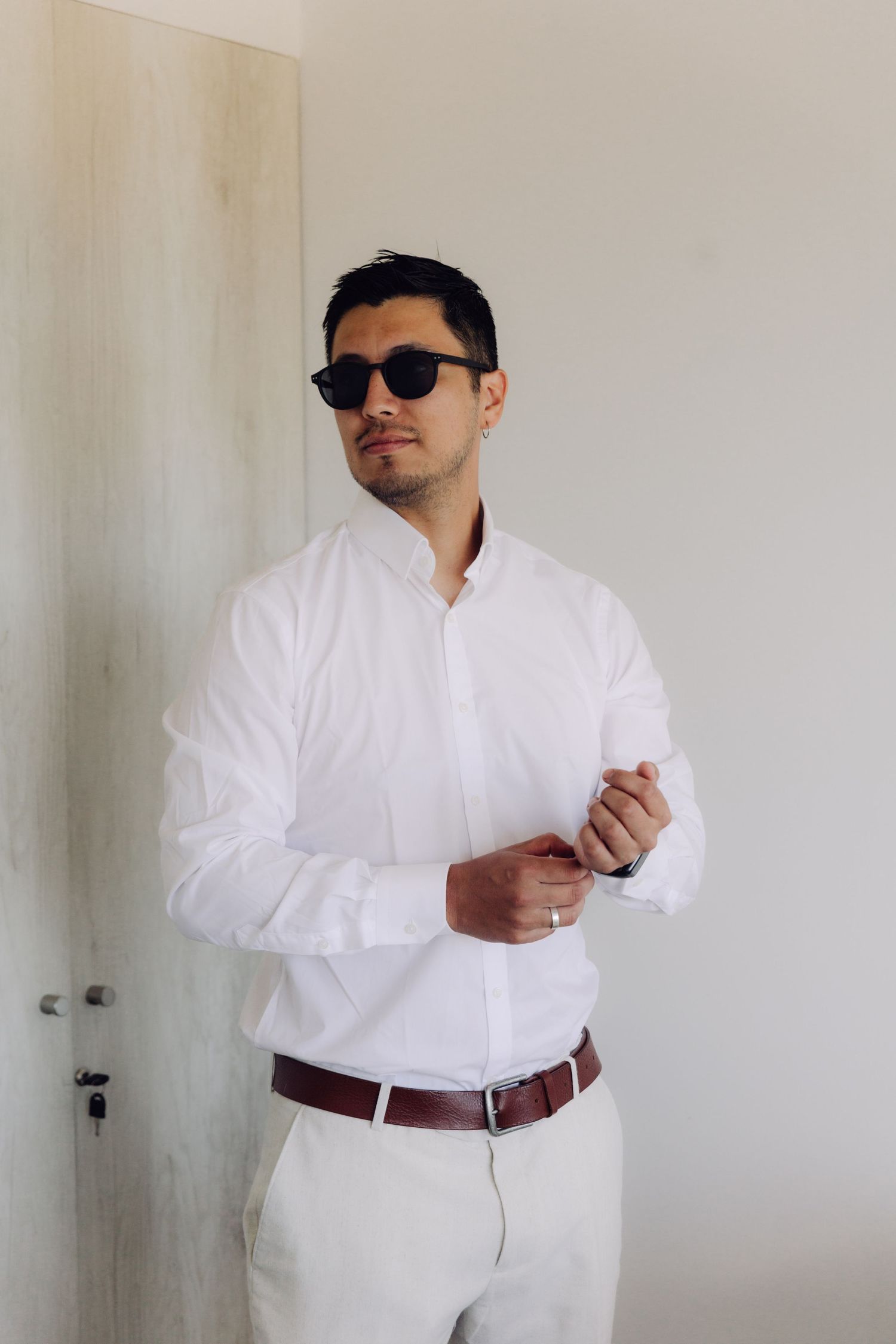 Elegant groom wearing white shirt, sunglasses, and brown belt standing by a door.