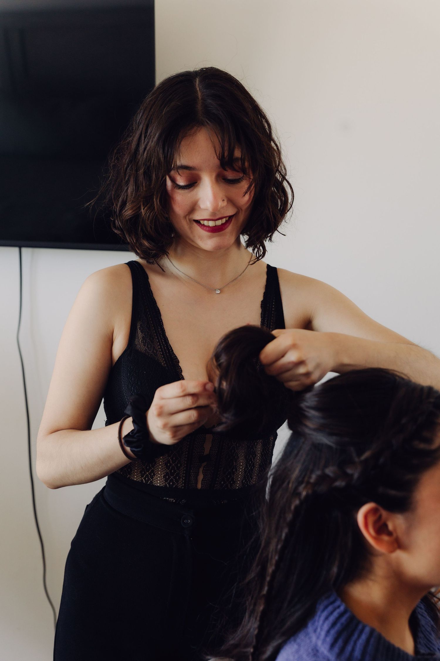 Hairstylist smiling while braiding dark-haired bride's hair during wedding preparation.
