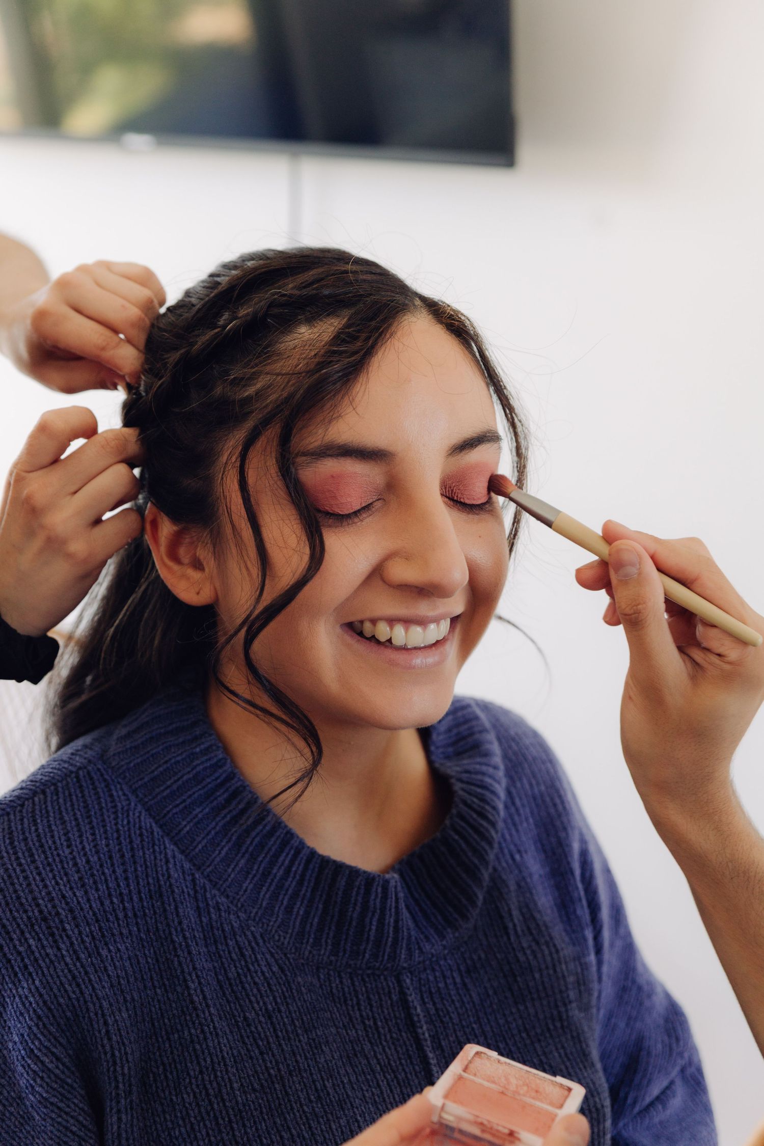 Bride smiling joyfully as makeup artist applies eyeshadow during bridal preparation.