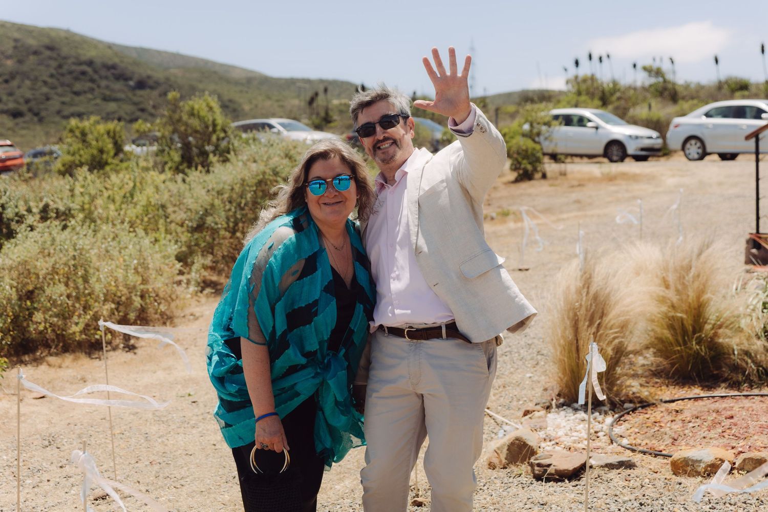 Smiling couple posing outdoors at a wedding venue with mountains and cars in background.