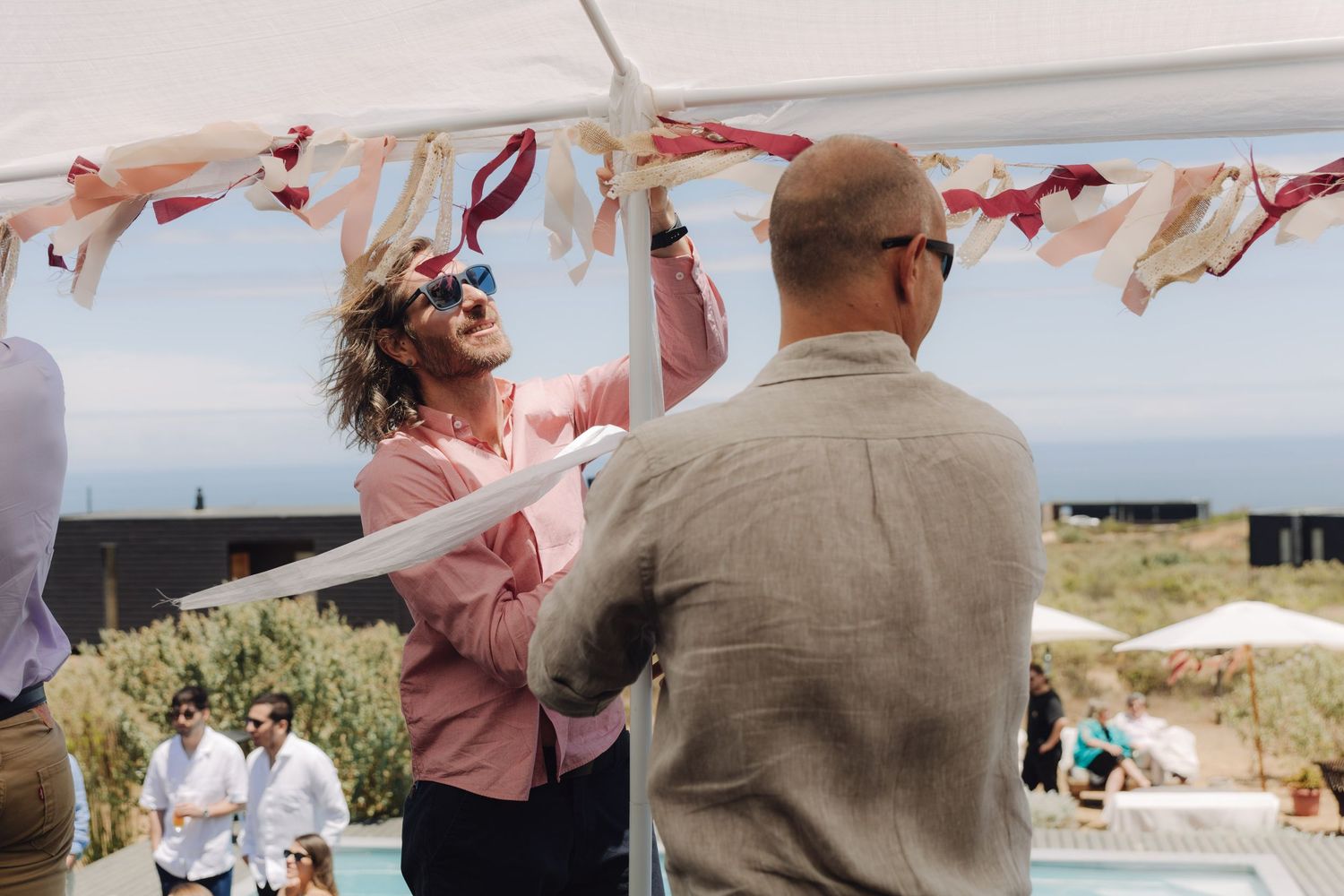 Man with curly hair arranging colorful ribbon decorations under a tent at an outdoor wedding celebration.