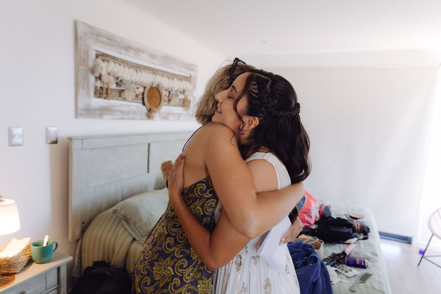 Two women sharing an emotional embrace and kiss in a bright white bedroom during wedding preparations.