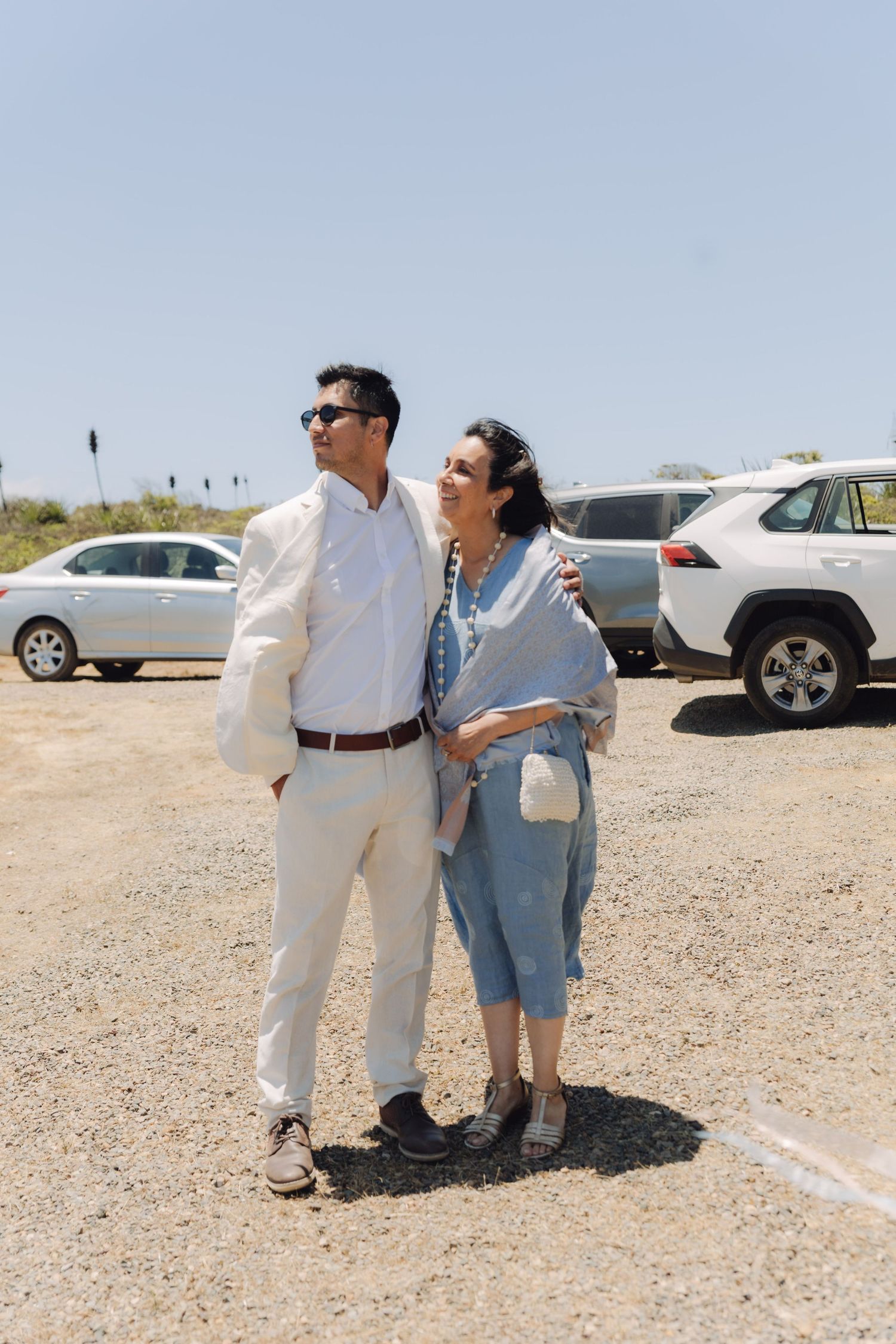 Elegant couple in formal attire standing together in a sunny outdoor parking area at a wedding.