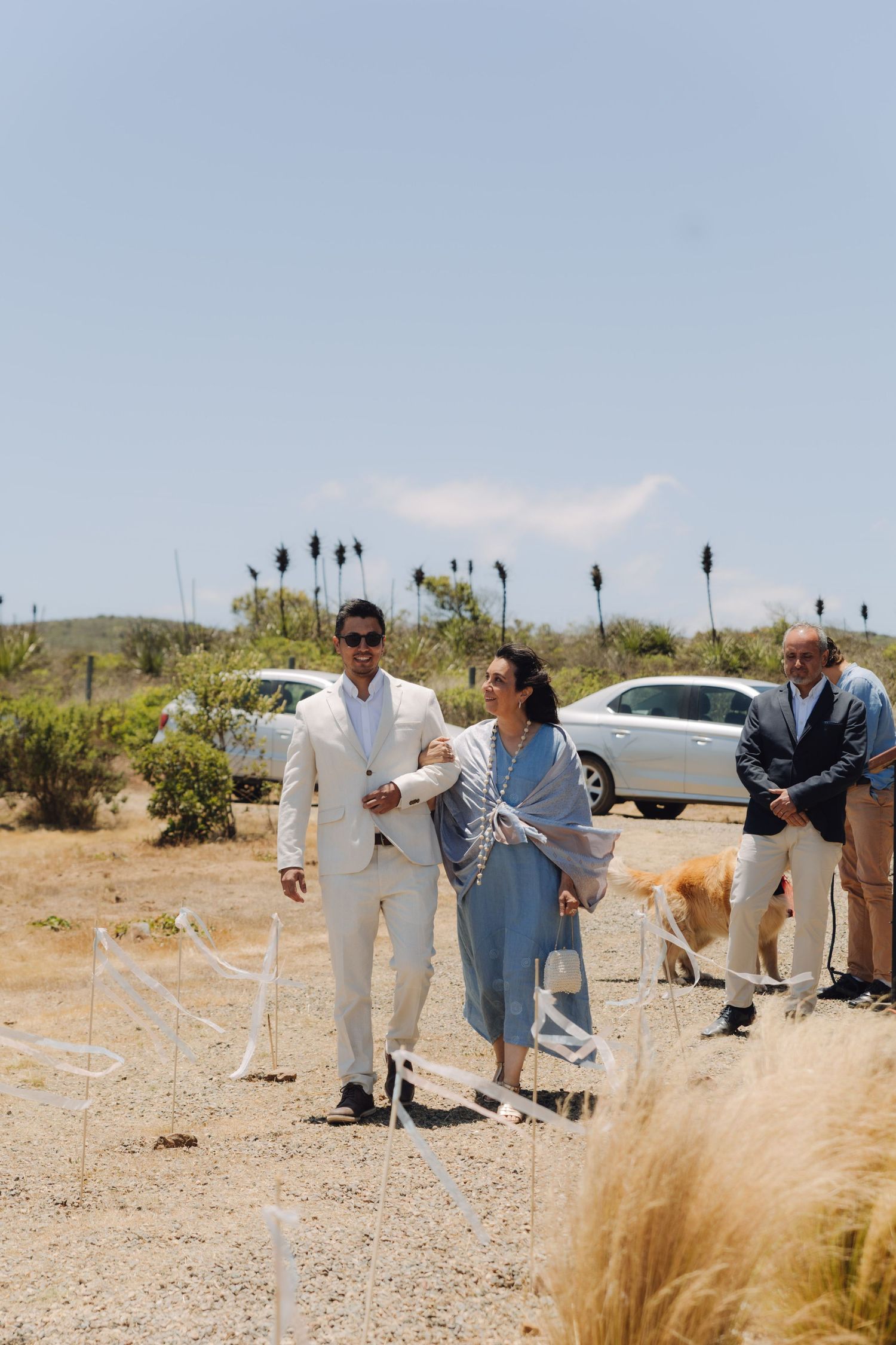 Groom in white suit walking down the aisle with a woman in blue at an outdoor desert wedding.