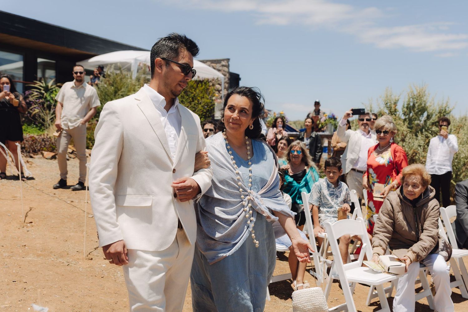 Groom in white suit escorting a woman in blue dress down the wedding aisle past seated guests.